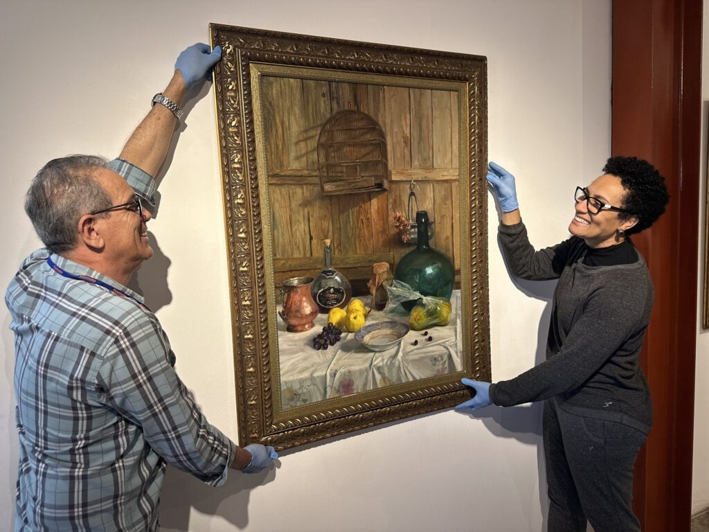 A imagem mostra duas pessoas, um homem e uma mulher, pendurando cuidadosamente um quadro em uma parede branca, provavelmente em uma galeria, museu ou exposição de arte. As Pessoas: Ambos estão usando luvas de proteção azuis (comuns para manusear obras de arte) para não danificar o quadro. O homem, à esquerda, usa óculos e uma camisa xadrez de tons azulados. A mulher, à direita, também usa óculos e está vestindo uma blusa cinza de gola alta. O Quadro: É uma pintura a óleo com uma moldura dourada trabalhada. O tema da pintura é uma natureza-morta clássica, retratando uma mesa com frutas (limões, uvas), uma garrafa grande de vidro verde, uma garrafa menor, uma jarra de metal e outros objetos, com uma gaiola ao fundo sobre uma parede de madeira. O Ambiente: A parede é branca e lisa, típica de espaços de exposição, o que ajuda a destacar a obra de arte. Parece ser um momento de montagem de uma exposição, feito com bastante cuidado e profissionalismo.