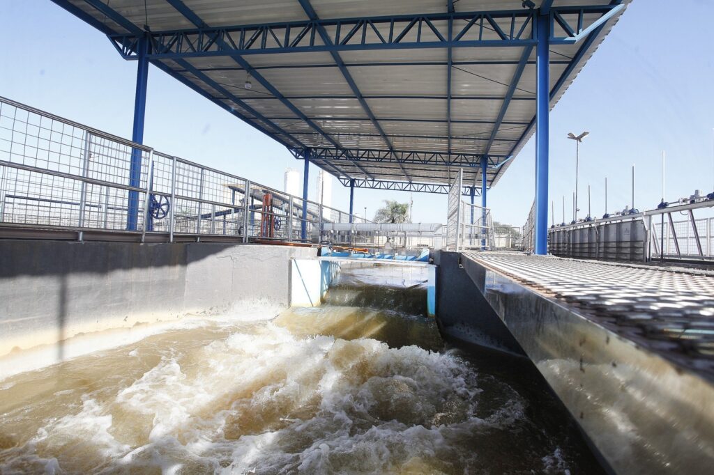 Esta imagem mostra uma estação de tratamento de água ou esgoto em pleno funcionamento. Estrutura: O local é coberto por uma estrutura metálica alta, com colunas azuis, que protege os tanques de tratamento. Processo: Em primeiro plano, é possível ver a água em movimento turbulento, passando por um canal de concreto que faz parte do sistema de filtragem ou oxigenação. A água apresenta uma coloração marrom, característica comum em certas etapas desse tipo de tratamento. Detalhes: Há passarelas metálicas gradeadas nas laterais para permitir o acesso e circulação dos operadores, com guarda-corpos de segurança e grades de proteção ao redor de toda a estrutura. O dia está ensolarado e com céu limpo, iluminando bem as instalações industriais.