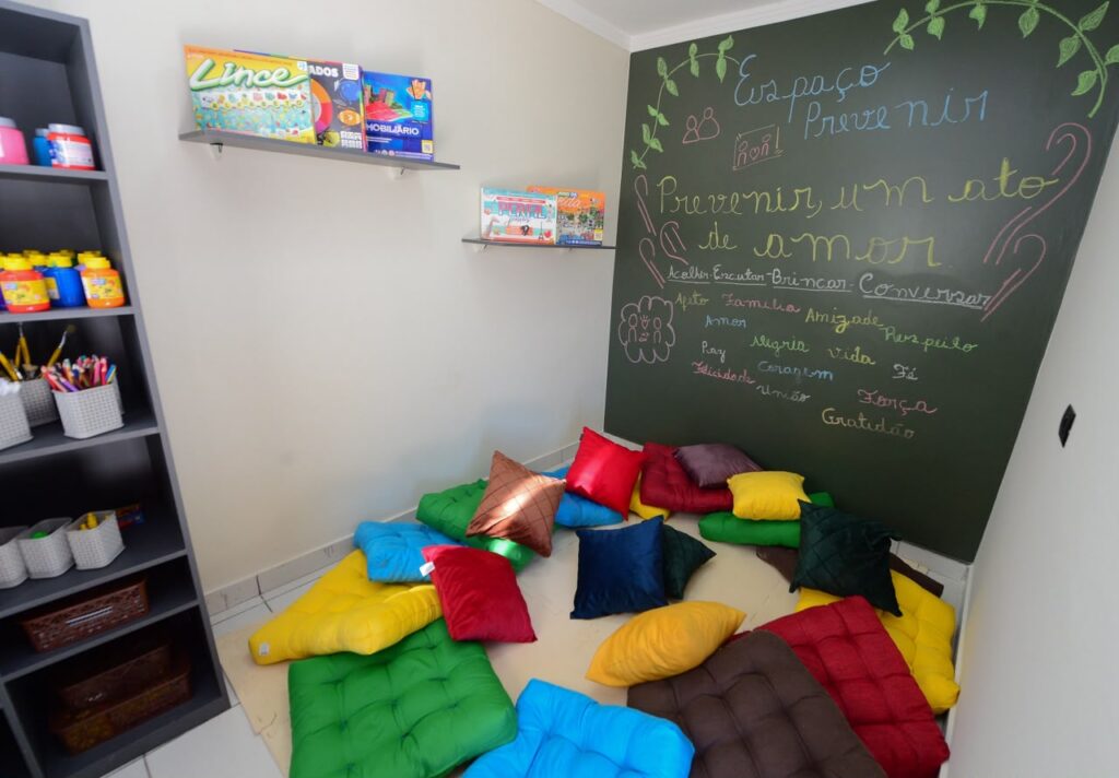 A imagem mostra uma sala de acolhimento com almofadas coloridas no chão, materiais lúdicos nas prateleiras e uma lousa com mensagens sobre prevenção, amor e convivência.