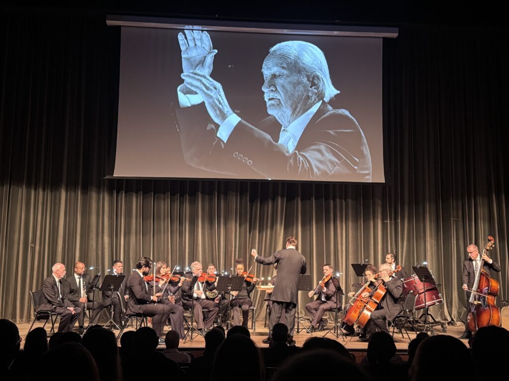 Esta é uma cena emocionante de uma apresentação de uma orquestra em um palco. Aqui estão os detalhes: O Telão: Acima do palco, há uma grande projeção em preto e branco de um maestro regendo, com uma expressão intensa e as mãos levantadas, o que parece ser uma homenagem ou uma participação especial virtual. A Orquestra: No palco, um grupo de músicos, todos vestidos de terno preto, está tocando seus instrumentos. Podemos ver diversos violinos, violoncelos e um contrabaixo à direita. O Maestro: De costas para a plateia, um maestro (também de terno preto) rege o grupo, criando uma conexão direta entre a música ao vivo e a imagem projetada acima. O Ambiente: O palco é decorado com cortinas pesadas e escuras ao fundo, dando um ar clássico e solene à apresentação. A plateia está sentada no escuro em primeiro plano, observando atentamente.