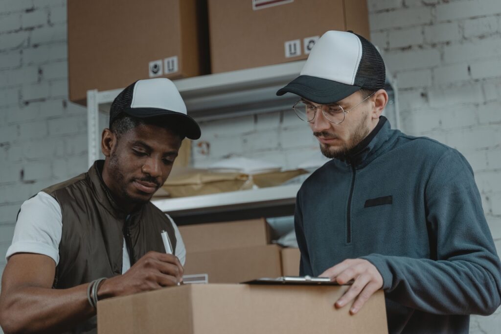 Foto de dois trabalhadores embalando e conferindo encomendas em um ambiente tipo depósito ou centro de distribuição. Detalhes observados: Dois homens lado a lado, ambos usando bonés tipo trucker com frente branca e aba/parte traseira preta. O homem à esquerda é mais escuro de pele, veste camiseta branca com colete sem mangas verde-oliva; segura uma caneta e parece assinar ou escrever algo sobre a caixa. O homem à direita usa óculos de armação fina, casaco cinza-azulado com zíper curto no peito e segura uma prancheta apoiada na caixa, conferindo informações. Há várias caixas de papelão empilhadas ao fundo, prateleiras metálicas e pacotes envelopados — ambiente organizado, típico de logística/armazenagem. Parede de tijolos brancos ao fundo, iluminação suave e neutra, sem sombras duras — cena com aspecto profissional e calmo. A caixa em primeiro plano é grande; a interação sugere conferência de itens, checklist ou preparação para expedição.