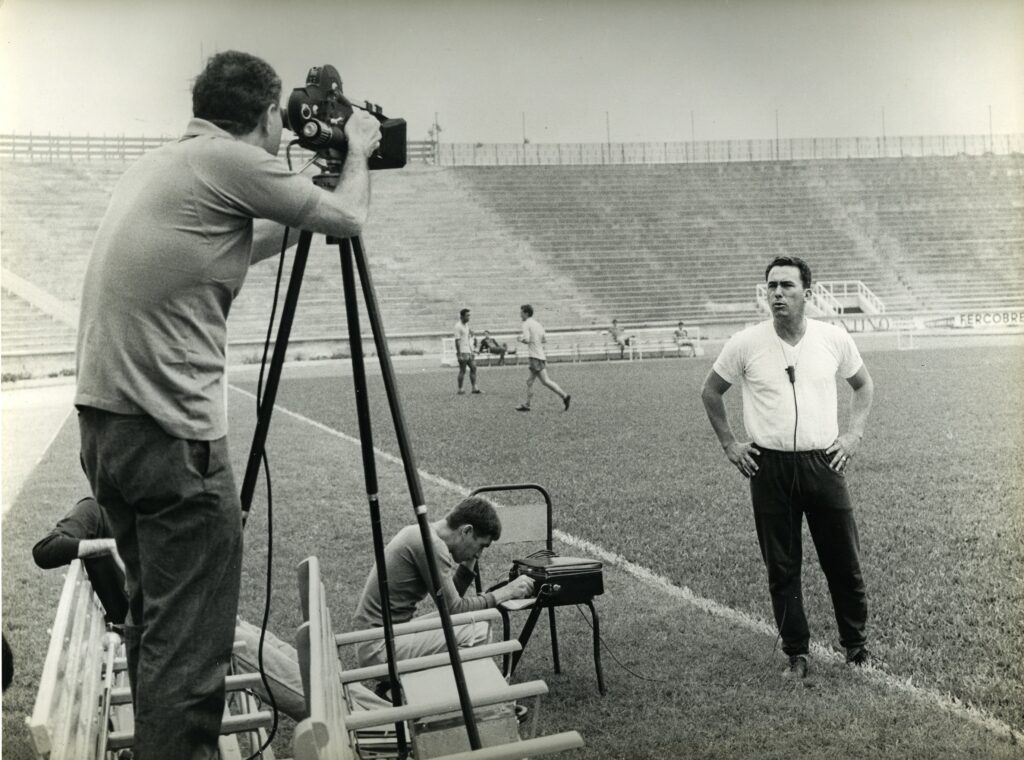 Foto em preto e branco, com aparência antiga. Cena num estádio vazio, provavelmente durante um treino. À esquerda, um cinegrafista de costas opera uma câmera grande montada num tripé. A câmera e os equipamentos parecem de época. No centro-direita, um homem em pé, de frente, com as mãos na cintura e apito pendurado no pescoço — parece um treinador ou instrutor, falando ou posando para a câmera. Ao fundo, alguns jogadores caminham ou conversam no gramado, vestindo roupas esportivas simples. Na parte inferior, outro homem sentado mexe num equipamento — possivelmente controlando a filmagem ou áudio. As arquibancadas estão vazias e dominam o plano de fundo, reforçando a sensação de espaço amplo e isolamento. Iluminação natural, clima neutro; o conjunto transmite um tom documental ou de registro de treino, com sensação de década passada (décadas de 1960–1980) por causa dos aparelhos e do estilo da imagem.