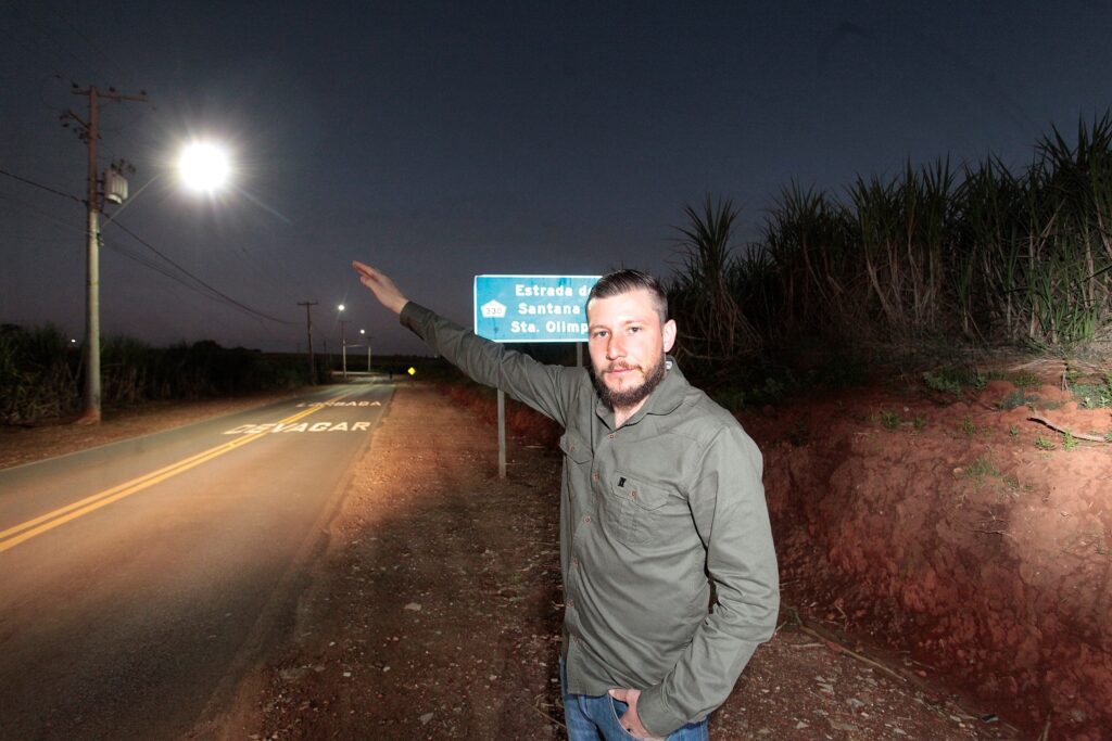 Homem em primeiro plano, posicionado à direita, parado na beira de uma estrada à noite; ele olha para a câmera e estende o braço direito apontando para cima/à esquerda. Cena iluminada por flash e por postes de iluminação pública; céu escuro de crepúsculo/noite. Pessoa (não identificável) Homem adulto, barba curta, cabelo penteado para trás; veste camisa/camperinha verde-oliva e jeans, com a mão esquerda no bolso. Expressão neutra/leveserena; não é possível identificar a pessoa. Ação e objetos Ele aponta na direção de um poste de luz muito brilhante e de uma placa de sinalização azul ao fundo. Placa parcialmente legível com texto parecido com “Estrada de Santana / Sta. Olímpia”. No asfalto, pintura de advertência “DEVAGAR” visível próximo ao primeiro plano. Ambiente e infraestrutura Estrada de pista dupla com faixas amarelas centrais; acostamento de terra batida. Postes e fiação elétrica ao longo da via; um poste próximo com transformador e luminária intensa. Laterais da estrada com cultivo alto (parece canavial) à direita e também à esquerda em distância. Talude de terra vermelha na margem direita, com vegetação rasteira.