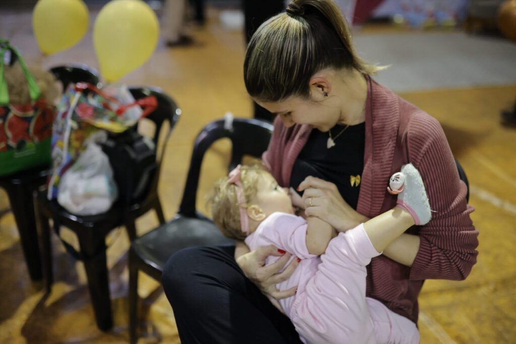 Esta é uma cena terna e íntima de uma mãe amamentando seu bebê. As pessoas: A mãe está sentada em uma cadeira preta, sorrindo gentilmente enquanto olha para o filho(a). Ela veste uma blusa preta, um cardigã rosa antigo e tem o cabelo preso em um rabo de cavalo. O bebê, que usa uma roupa rosa, uma faixa de cabelo com um laço e meias com detalhes fofos, está deitado confortavelmente no colo dela, segurando a mão da mãe. O ambiente: O cenário parece ser um evento ou salão, com piso de madeira, cadeiras pretas ao fundo e balões amarelos, sugerindo uma celebração ou reunião familiar. A iluminação é suave e focada nas duas, criando um momento de conexão muito bonito e sereno.
