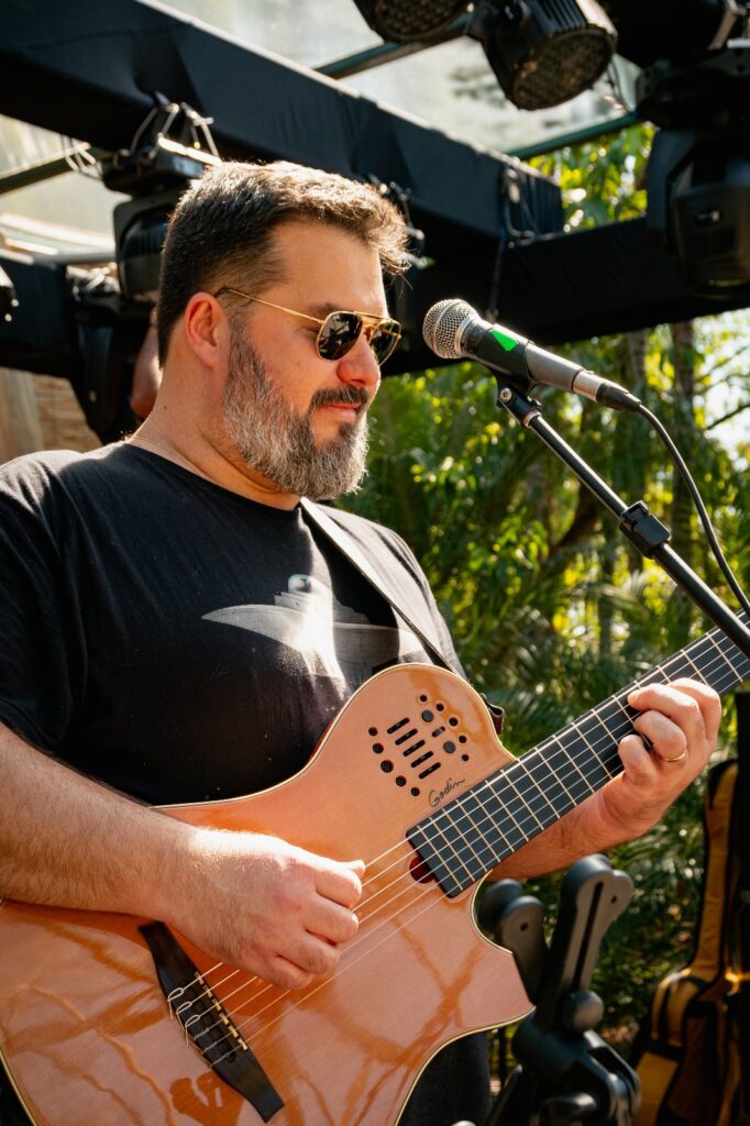 Esta foto captura um momento de um músico se apresentando ao vivo. O Músico: Um homem de meia-idade, com barba grisalha e óculos escuros estilo aviador, está tocando um violão. Ele usa uma camiseta preta com uma estampa que parece ser um disco voador ou objeto espacial. O Instrumento: Ele toca um violão de nylon da marca Godin (modelo Multiac), característico pelo seu design mais fino e moderno, sem a boca tradicional no centro da caixa de ressonância. O Ambiente: A cena acontece ao ar livre ou em um espaço com luz natural, rodeado por folhagens verdes ao fundo. Equipamento: Há um microfone posicionado à sua frente e é possível ver equipamentos de iluminação de palco (canhões de luz) na parte superior do enquadramento, indicando que é um cenário de show ou evento musical profissional. O músico parece estar focado e concentrado na execução da música.