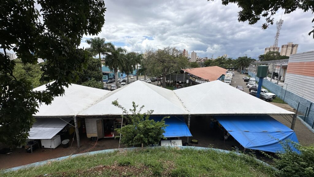 A imagem mostra uma vista aérea ou de um ponto elevado de uma área externa com três tendas brancas grandes conectadas, cobrindo uma área de comércio ou eventos. Abaixo das tendas, há diversas estruturas temporárias, como trailers e balcões, alguns protegidos por lonas azuis e brancas. Ao redor da área das tendas, vê-se um estacionamento com vários carros, além de árvores, postes de iluminação e vegetação rasteira em primeiro plano. Ao fundo, nota-se uma área urbana com prédios e um céu nublado, indicando um dia com nuvens carregadas.