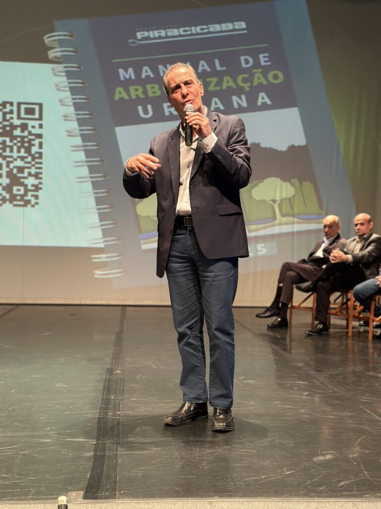 Foto de um evento no palco com um orador em primeiro plano falando ao microfone. Observações: Pessoa em destaque: um homem adulto de pele clara, cabelos curtos e grisalhos, vestido com paletó escuro, camisa clara e jeans; segura um microfone com a mão direita e faz um gesto com a mão esquerda, em atitude de fala explicativa. Cenário: palco com projeção grande ao fundo mostrando a capa de um documento/ manual — é possível ler parcialmente "MANUAL DE ARBORIZAÇÃO URBANA" e o logotipo/identificação "PIRACICABA", sugerindo tema ligado a arborização ou gestão urbana. À esquerda da projeção aparece um QR code ampliado. Público/participantes: ao fundo, sentados em cadeiras no palco, ao menos dois homens (aparentando serem membros da mesa ou painel), observando; estão vestidos de forma formal. Iluminação e ambiente: luz de palco direcionada ao orador, com boa definição de cor e sombras suaves; chão brilhante do palco reflete iluminação. Ambiente parece ser auditório ou teatro em evento institucional. Composição e sensação: enquadramento vertical que valoriza o orador em movimento; imagem transmite formalidade e discurso público sobre políticas ou práticas urbanas (arboretação).