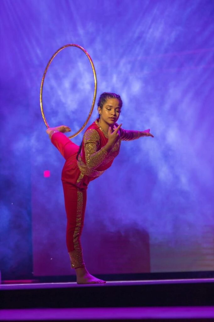 Esta foto captura um momento elegante e artístico de uma apresentação de dança ou ginástica. A Pessoa: Uma jovem com o cabelo preso em tranças está realizando uma pose de equilíbrio em um pé só. Ela demonstra muita concentração e graciosidade. O Figurino: Ela veste um conjunto nas cores vermelho e dourado, que brilha sob as luzes, com detalhes em lantejoulas ou tecido metalizado nas mangas e nas laterais das calças. O Movimento: Ela está equilibrando um bambolê dourado no ar, logo acima de suas costas, enquanto mantém uma perna elevada para trás, criando uma silhueta dinâmica e bonita. O Ambiente: O cenário é um palco com iluminação cênica vibrante, com tons fortes de roxo e azul, e um efeito de fumaça ao fundo que dá um toque dramático e profissional à apresentação. É uma imagem que transmite muita disciplina, talento e impacto visual!