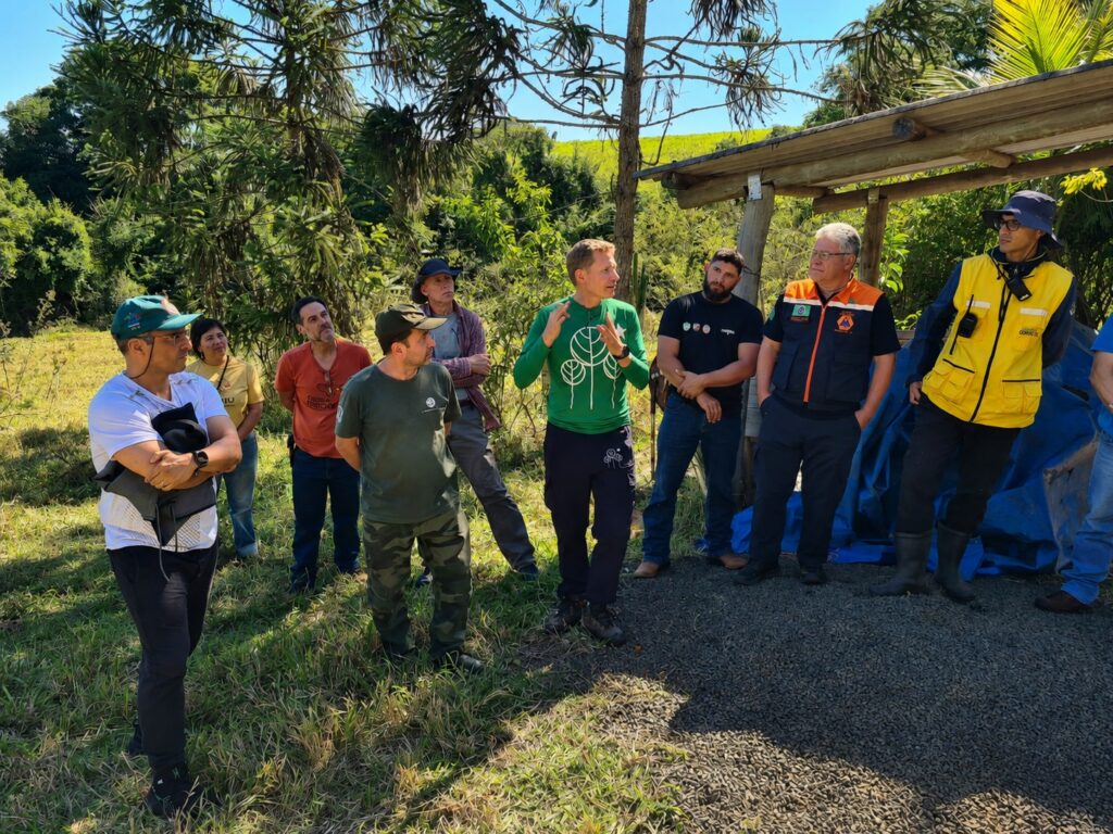 A foto registra um grupo de pessoas em um encontro ao ar livre, possivelmente uma atividade de campo, treinamento ou visita técnica. As Pessoas: Há um grupo de cerca de oito pessoas em pé. No centro, um homem veste uma camiseta verde de manga comprida com um desenho de árvores brancas e gesticula enquanto fala, parecendo estar liderando ou explicando algo para o grupo. À direita, há um homem vestindo um uniforme laranja e azul marinho, com um colete de identificação e aparência de autoridade (como Defesa Civil ou órgão público). Ao lado dele, outro homem usa um colete amarelo de alta visibilidade sobre uma jaqueta escura, também com equipamento. As outras pessoas ao redor vestem roupas casuais (camisetas, calças jeans/camufladas, bonés) e observam atentamente o orador. O Ambiente: Estão em uma área com vegetação rasteira e algumas árvores, incluindo um exemplar de araucária ao fundo. Há uma estrutura de madeira simples (um telhado ou abrigo) à direita, protegendo um monte de brita ou cascalho sob uma lona azul. Clima: O dia está ensolarado e a iluminação é forte, criando sombras naturais no chão. O ambiente transmite a sensação de um trabalho de campo ou vistoria em área rural ou de preservação.
