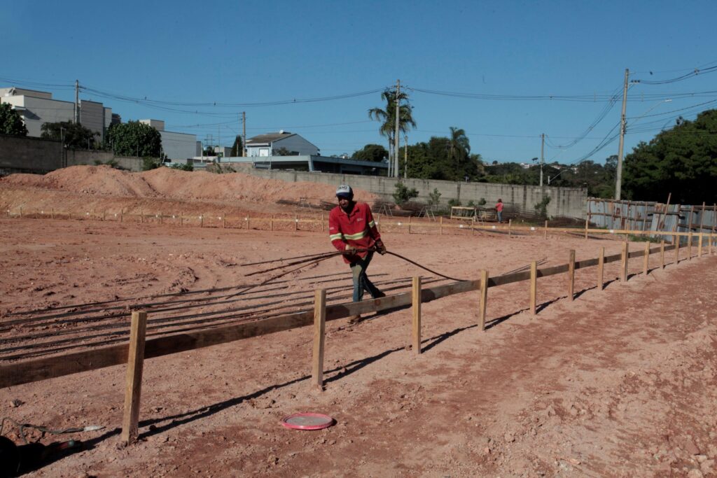 Foto de um canteiro de obras ao ar livre em dia ensolarado. Cena geral: terreno amplo de terra avermelhada nivelada, com marcadores de madeira formando divisões para fundação. Pessoas: um trabalhador em primeiro plano, veste camisa vermelha com faixas refletivas e boné, puxando vergalhões/armaduras; ao fundo, outro trabalhador perto do muro. Materiais e equipamentos: vergalhões dispostos paralelos no chão, estacas e tábuas de forma, pequenas pilhas de terra ao fundo; tampinha ou tampa plástica rosa no chão à frente. Ambiente: muro de contenção e construções residenciais ao fundo; postes e cabos elétricos atravessam a cena; vegetação e algumas árvores além do muro. Iluminação e clima: céu azul sem nuvens visíveis, luz forte criando sombras curtas — parece manhã ou começo da tarde. Observações: área aparenta estar na fase inicial de demarcação e preparação de fundações.