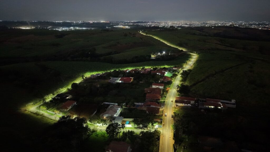 Foto aérea noturna mostrando um trecho semi‑rural: um bairro/aglomerado de casas iluminadas próximo a uma estrada que corta campos e morros até um conjunto de luzes urbanas ao longe. Perspectiva em plongée que guia o olhar pela via iluminada desde o primeiro plano até o horizonte. Estrada e iluminação Estrada asfaltada bem visível à direita, com postes de iluminação formando uma faixa contínua de luz amarelada. Outra via menor, mais à esquerda, também aparece iluminada por postes, contornando o condomínio/aglomerado de casas. As luzes do poste criam faixas brilhantes sobre o asfalto e refletem nas fachadas das casas, destacando o núcleo habitado no escuro. Casas e área construída Conjunto de residências com telhados vermelhos e quintais; algumas casas têm iluminação externa clara que evidencia muros, piscinas e áreas gramadas. Ruas internas e cercas são perceptíveis graças aos pontos de luz, formando um reticulado dentro da mancha urbana pequena. Vegetação e terreno Grandes áreas verdes e campos escuros ao redor das casas, com relevo ondulado e morros suaves que criam contraste entre zonas iluminadas e sombreadas. Trechos de mata/arbustos aparecem como massas escuras que separam a área urbana das luzes ao fundo. Fundo e horizonte No horizonte, extensa mancha de luzes urbanas indica uma cidade ou aglomerado maior distante; céu noturno levemente acinzentado sem estrelas visíveis devido ao brilho urbano. A estrada que parte do núcleo parece seguir em direção a esse ponto luminoso, reforçando sensação de ligação entre zona rural e cidade.