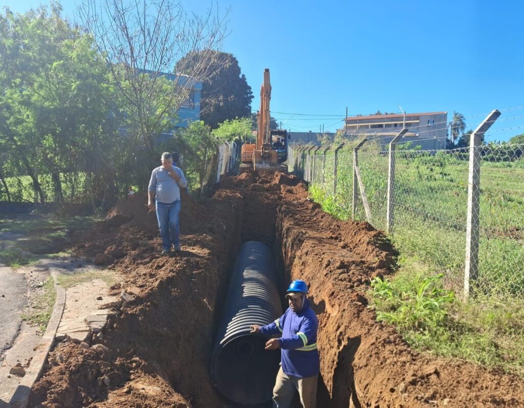 Imagem colorida de uma obra externa durante o dia, mostrando a instalação de uma grande tubulação em valeta. Cena geral: valeta longa e profunda escavada no solo ao longo de uma cerca de arame e postes de concreto; solo escuro e montes de terra à beira da vala. Equipamento: escavadeira laranja posicionada ao fundo da vala, com o braço voltado para a frente, aparentemente em operação. Tubulação: grande tubo corrugado preto já assentado no fundo da vala, ocupando boa parte do comprimento visível. Pessoas: dois trabalhadores visíveis — um dentro da vala, usando capacete azul e roupa com faixas refletivas, segurando ou orientando o tubo; outro homem em cima da borda da vala, com roupa casual, olhando para baixo. Não é possível identificar quem são. Ambiente: ao lado direito há um terreno gramado cercado; ao lado esquerdo árvores e vegetação alta que fazem sombra parcial. Ao fundo, construções residenciais e céu azul claro, sem nuvens. Iluminação e clima: luz solar forte, sombras nítidas; aparenta ser manhã ou início de tarde com clima claro e seco. Impressão: trabalho organizado de infraestrutura (provavelmente drenagem ou escoamento pluvial), com movimentação de terra e assentamento de tubulação em andamento.