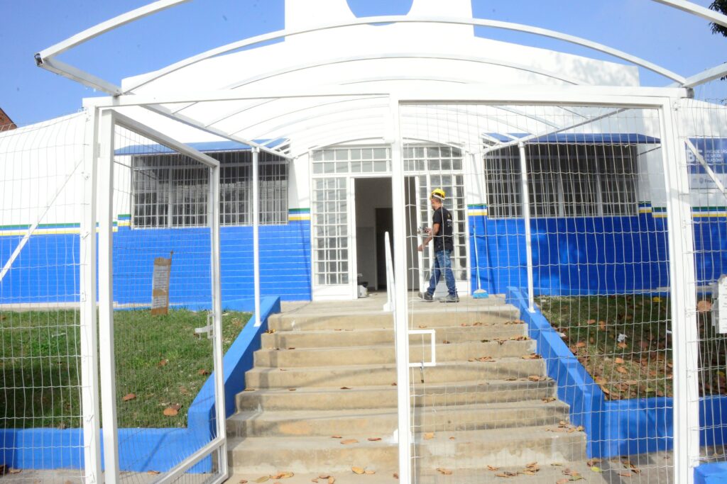 Entrada de um prédio institucional vista de frente, com portão e cobertura metálica branca formando um corredor até a porta principal. Estrutura: portão gradeado branco aberto; cobertura curva metálica branca sobre o caminho; escada de concreto com corrimãos pintados de azul que leva à entrada. Fachada: parede pintada em branco e azul; faixa com listras verde, amarela e branca acima da parte azul. Pessoas/atividades: um homem usando capacete amarelo e roupa escura caminha próximo à porta — parece trabalho ou manutenção (não faço identificação). Detalhes no entorno: grades nas janelas, área gramada nas laterais com algumas folhas secas espalhadas, uma vassoura encostada perto da entrada. Ambiente/iluminação: dia claro com céu azul, iluminação natural forte e sombras suaves.