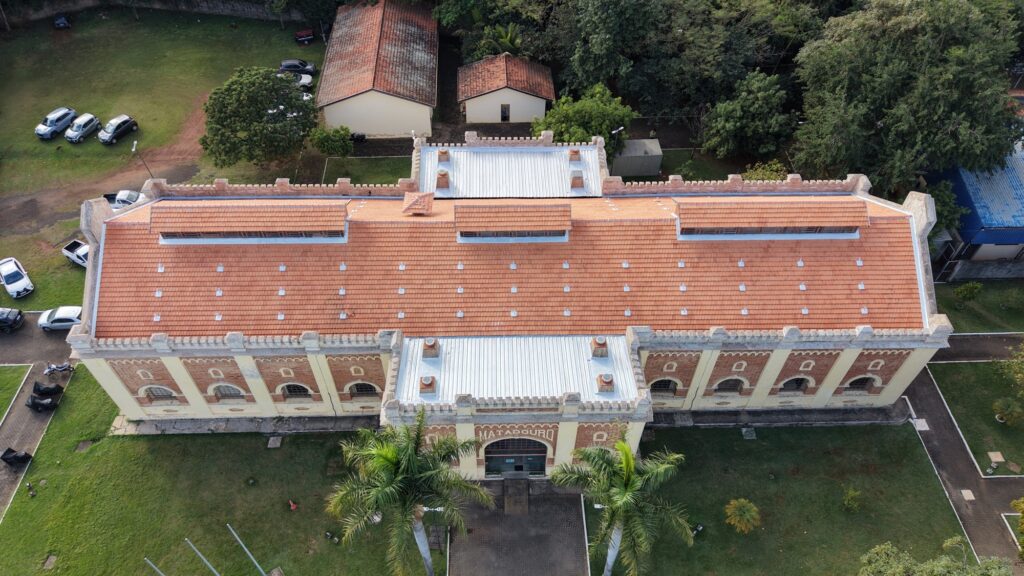 Esta foto oferece uma vista aérea impressionante do edifício que você enviou, destacando perfeitamente a sua simetria e arquitetura: O Edifício: É uma estrutura retangular longa com telhado de duas águas em telhas cerâmicas avermelhadas. O corpo central, onde fica a entrada principal, se projeta para a frente com um telhado metálico e um acabamento que ostenta a inscrição "MATADOURO" logo acima da porta de vidro. Detalhes do Telhado: Podemos notar várias pequenas claraboias ou lanternins ao longo do telhado, fundamentais para a ventilação e iluminação natural desse tipo de construção antiga. Há também diversos grampos metálicos distribuídos uniformemente pelo telhado. Entorno: O prédio está inserido em uma área verde muito bem cuidada. À esquerda, há um estacionamento organizado com vários carros. Atrás, notamos construções menores e mais simples com telhados marrons. Dois coqueiros altos emolduram a entrada principal, dando um toque tropical à cena. À direita e ao fundo, há bastante vegetação, incluindo árvores densas. É um registro muito claro de como a conservação desse prédio histórico combina com a organização funcional do pátio ao redor.