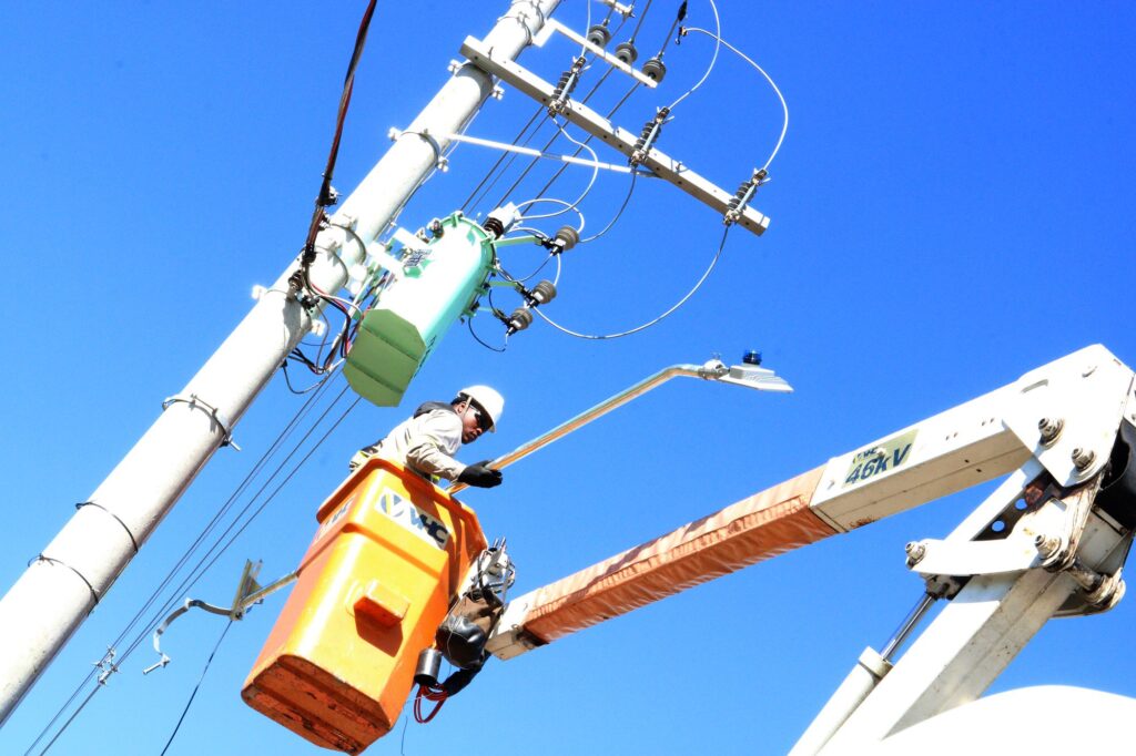 A imagem mostra um trabalhador de manutenção elétrica realizando serviços em um poste de energia. Ele está dentro de um cesto aéreo laranja acoplado a um caminhão com braço hidráulico, manipulando uma luminária pública. O profissional usa equipamentos de proteção individual, como capacete e luvas, enquanto trabalha próximo a fios e transformadores. O céu azul intenso ao fundo destaca o ambiente externo e o clima ensolarado.