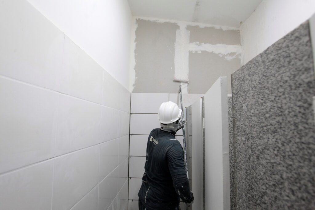 Foto de um trabalhador pintando uma parede em um ambiente interno, possivelmente um banheiro em reforma. Pessoa: um trabalhador de costas usando capacete branco e roupa escura com respingos de tinta. Ação: ele segura um rolo de pintura em extensão e aplica tinta clara em uma parede alta; já há uma faixa vertical e marcas de massa. Ambiente: paredes azulejadas brancas à esquerda e divisórias de banheiro em granito ou material similar à direita; chão e parte inferior fora de vista. Detalhes: iluminação branca e ambiente estreito; parede superior sem azulejos mostrando reboco e áreas em fase de acabamento.