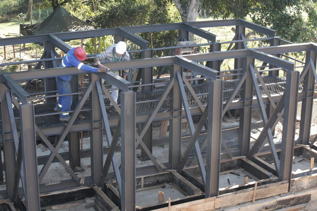 Uma estrutura metálica em construção vista de cima, com dois operários medindo e alinhando perfis de aço. Detalhes: Vigas e pilares de aço cinza-escuro formando uma grelha retangular apoiada sobre pilares e contraventamentos em X. Dois trabalhadores no topo: um com capacete vermelho e camiseta azul, outro com capacete branco e casaco listrado; ambos manipulando um nível ou régua de medição. Plataforma metálica tipo grelha sob as vigas, escada de mão encostada ao fundo. Ao redor, vegetação e árvores, indicando canteiro de obras ao ar livre; há também formas de madeira e fundações de concreto na base. Iluminação natural forte, sombras suaves das árvores sobre a estrutura. Aparente fase de montagem/estruturação inicial antes do fechamento ou revestimento.