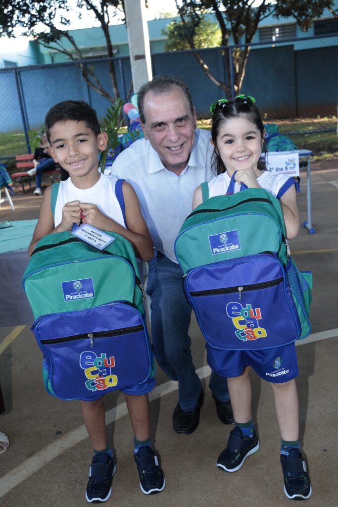 Foto de três pessoas sorrindo em um evento escolar ao ar livre. Pessoas: dois crianças em primeiro plano (um menino à esquerda e uma menina à direita) segurando mochilas escolares grandes; um adulto (homem) agachado ao centro, entre as crianças, sorrindo para a câmera. Idade aparente e expressão: crianças em idade escolar inicial (próximo ao ensino fundamental), ambas sorrindo; o adulto também sorri de forma cordial. Roupa e objetos: crianças usam uniforme azul e branco; mochilas predominantemente verdes e azuis com o selo "Prefeitura de Piracicaba" e a palavra "educação" estampada; a menina tem laços verdes no cabelo. Cenário: área externa coberta (possivelmente quadra ou pátio escolar) com piso de concreto; ao fundo há placas, grade e árvores que indicam ambiente escolar. Iluminação e clima: luz natural, dia claro; atmosfera parece festiva ou de entrega de material escolar.