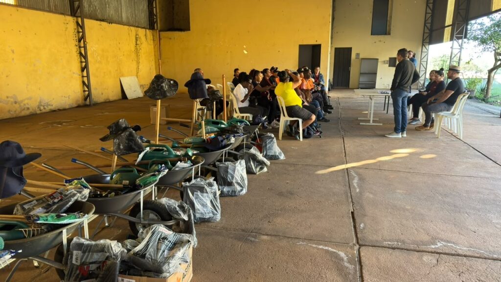 Uma reunião ou entrega de insumos num galpão/quadra coberta com parede amarela — pessoas sentadas à direita recebem instruções enquanto, à esquerda, uma fileira de carrinhos de mão e equipamentos está organizada para distribuição. Ambiente: espaço amplo e coberto com piso de concreto e parede lateral pintada de amarelo gasto; estrutura metálica visível e portas ao fundo; iluminação natural entrando pelas aberturas à direita. Pessoas: cerca de 15–20 pessoas sentadas em cadeiras plásticas formando duas filas, a maioria de frente para três pessoas em pé/assentadas que parecem conduzir a atividade. Vestimentas casuais: algumas pessoas com coletes laranja, outras com camisas escuras ou camiseta amarela. Atividade: parece uma orientação, briefing ou entrega — pessoas prestam atenção aos condutores e algumas conversam entre si. Materiais organizados: ao longo do lado esquerdo há uma sequência de carrinhos de mão (carrinhos metálicos) cada um com ferramentas e itens embalados — sacos plásticos, pás/ancinhos/manuais (alças visíveis), além de chapéus sobre suportes de madeira. Vários pacotes ainda embalados no chão. Disposição: os equipamentos estão alinhados ordenadamente próximos ao primeiro plano, prontos para distribuição; as cadeiras e a mesa branca ao fundo sugerem organização do evento.