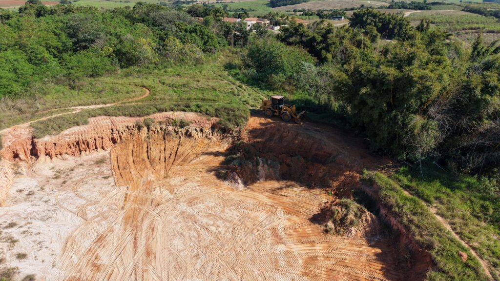 Vista aérea de uma área de escavação/aterro em terreno rural. Em primeiro plano e ocupando a maior parte da cena, há solo exposto de cor avermelhada e claras marcas de esteira de pneus formando padrões concêntricos — sinal de movimentação recente de máquinas. À esquerda, um escarpamento com camadas de solo e pequenas erosões revela a altura cortada do terreno. No canto direito do platô, há uma pá-carregadeira amarela estacionada sobre terra batida junto à borda, parcialmente sombreada pelas árvores. Ao redor da área escavada há vegetação densa: à direita um bosque com bambus e árvores altas; ao fundo, uma faixa de mata contínua que separa a cava de campos agrícolas e pequenas edificações rurais visíveis ao longe. Em níveis superiores da imagem vê‑se pasto e áreas cultivadas que se estendem até o horizonte. A iluminação sugere um dia claro e ensolarado, com sombras definidas das árvores. A composição transmite atividade de terraplenagem ou extração em um sítio rural, com contraste entre o solo nu e a vegetação intacta nas bordas.