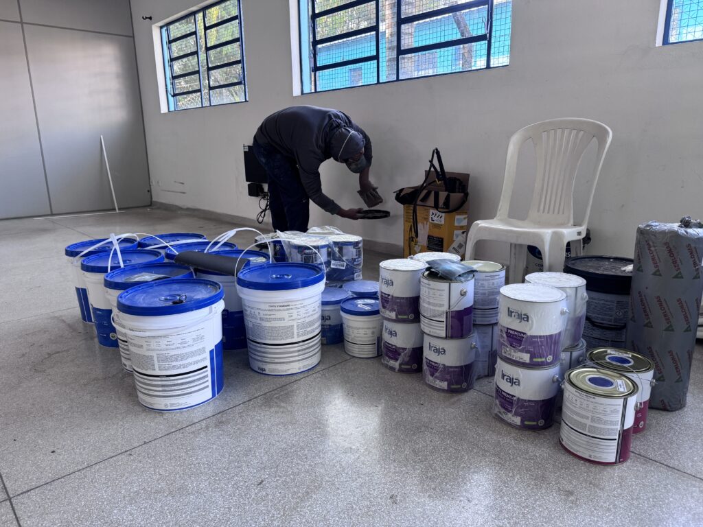 Sala interna clara com materiais de pintura organizados em primeiro plano e um trabalhador agachado perto da parede realizando preparo/inspeção. Composição geral: ambiente amplo com piso de granilite, paredes brancas e janelas altas com grades e venezianas azuis permitindo luz natural. Pessoa: um trabalhador vestido com moletom escuro, calça escura e boné/ touca, inclinado sobre a parede enquanto segura uma espátula/placa — parece executar acabamento ou checar algo na tomada/caixa embutida. Não faço identificação. Materiais: várias latas e baldes de tinta dispostos em pilhas e fileiras no chão — tampas azuis, rótulos visíveis de marcas; também rolo de pintura, lixas/espátulas e alguns recipientes menores. Equipamentos e caixas: caixa de papelão e alguns acessórios (cabos, ferramentas) junto à parede; saco de material (possivelmente forro ou isolamento) à direita. Mobiliário: cadeira plástica branca colocada ao lado, sem uso. Estado e ação: ambiente em fase de preparação ou reforma interna (produção/repintura). Materiais empilhados indicam trabalho em andamento. Iluminação e clima: dia claro com luz natural entrando pelas janelas, sombras suaves no piso.