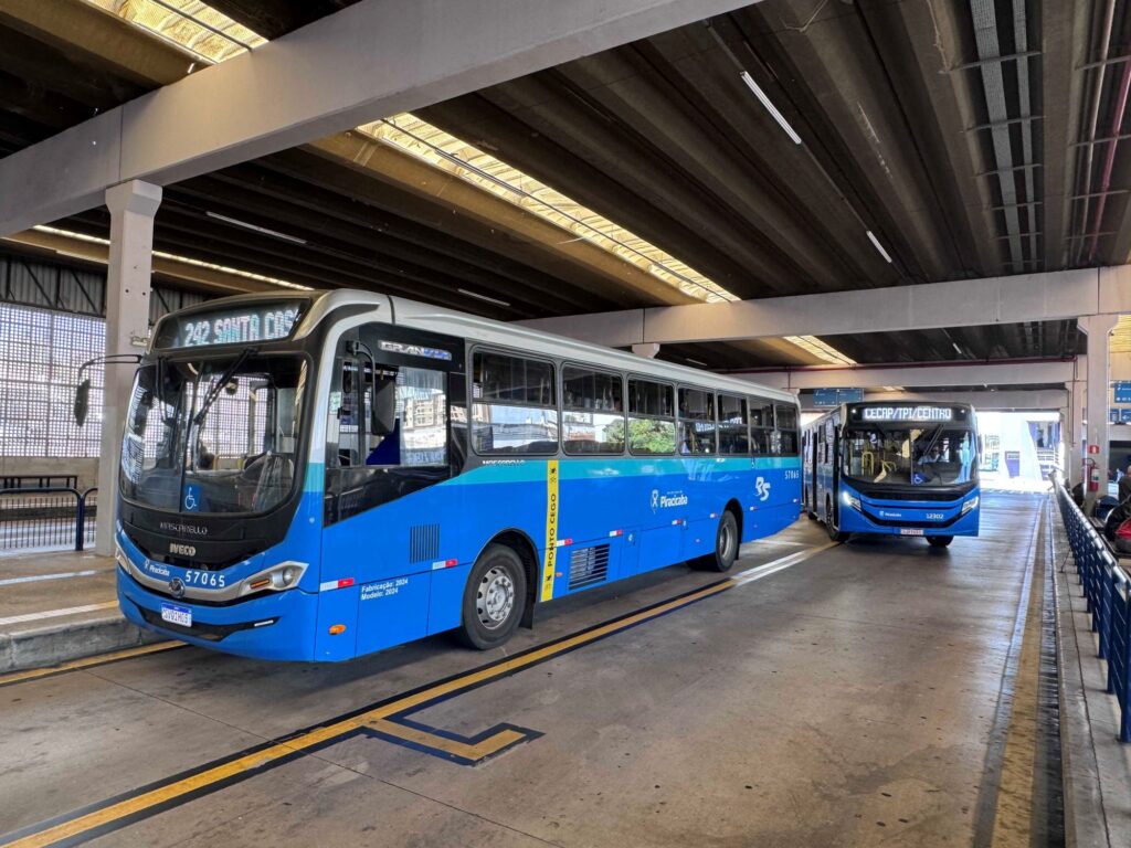 Esta imagem mostra um terminal de ônibus coberto com dois veículos da frota municipal de Piracicaba em destaque. Aqui estão os detalhes principais: Os ônibus: Ambos são modelos urbanos modernos, na cor azul predominante, com o brasão da Prefeitura de Piracicaba e acessibilidade para pessoas com deficiência. O ônibus em primeiro plano é da marca Iveco/Mascarello (modelo 2024), com o itinerário digital indicando "242 SANTA C...". O número da frota é "57065". O ônibus logo atrás exibe no itinerário "CECAP/TPI/CENTRO" e tem o número de frota "12302". O local: Trata-se de uma plataforma de embarque coberta com estrutura de concreto e vigas aparentes. O piso é de cimento, com faixas amarelas de sinalização. O ambiente: O terminal parece amplo, iluminado por luz natural vinda de aberturas no teto e laterais, e parece estar em um momento de movimentação tranquila.