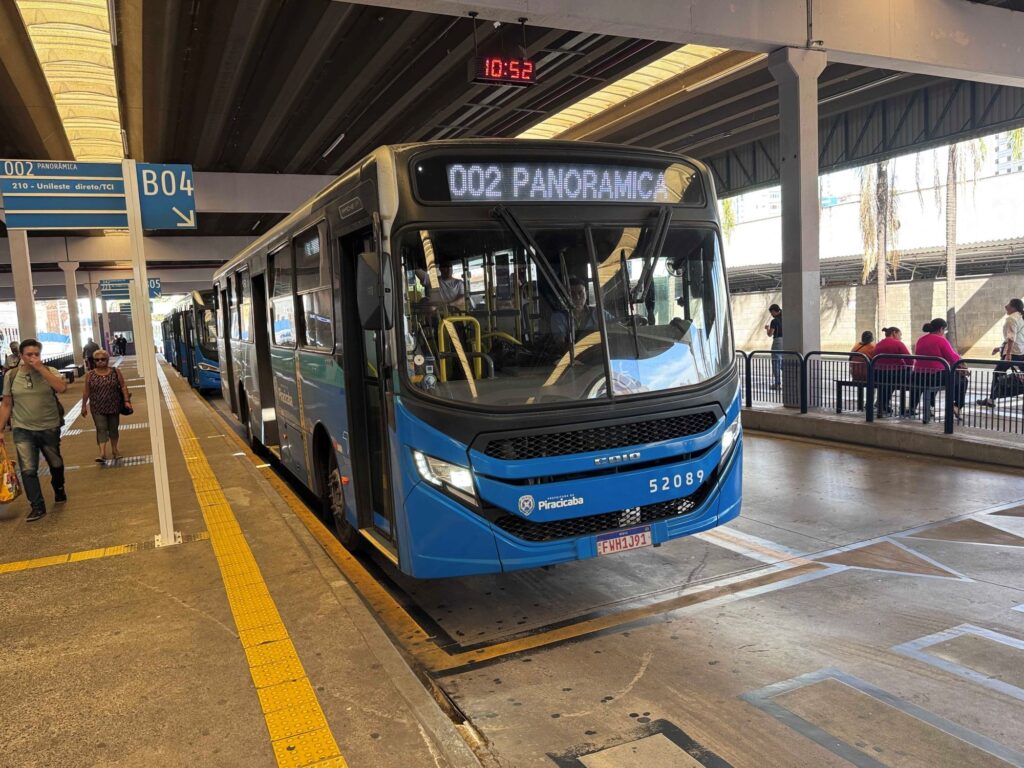 Essa é uma foto de um terminal de ônibus com um ônibus azul parado no ponto. Aqui estão os detalhes principais: O Ônibus: É um ônibus urbano moderno, na cor azul, com o itinerário digital indicando "002 PANORÂMICA". Ele exibe o brasão da "Prefeitura de Piracicaba" e o número de frota "52089". O Local: O ônibus está parado em uma plataforma de embarque coberta, identificada como "B04". Há um painel informando que o ônibus segue para "002 PANORÂMICA" e "210 - Unileste direto/TCI". Ambiente: Há passageiros andando pela plataforma e outros sentados em bancos próximos à grade de proteção à direita, aguardando ou descendo do transporte. Detalhes adicionais: O relógio digital suspenso na estrutura do terminal marca "10:52". O ambiente parece bem organizado e é um cenário típico de transporte público em uma cidade brasileira.