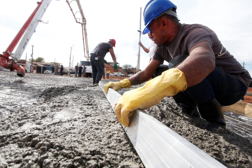 Um canteiro de obras visto em ângulo baixo e amplo, com foco em trabalhadores nivelando concreto recém-lançado. Detalhes observáveis: Cena principal: dois operários no primeiro plano trabalhando sobre uma laje de concreto úmido. Um deles, à direita, usa capacete azul, camiseta escura e luvas amarelas de borracha; está agachado e alisando o concreto com uma régua metálica longa. Outro, ao centro, com capacete vermelho, está de pé auxiliando a espalhar o material. Material e ação: grande volume de concreto fresco sendo adicionado e nivelado sobre uma armação metálica (malha de vergalhões) visível sob o concreto. A superfície está molhada e texturizada, ainda por acabamento. Equipamento: braço de bomba de concreto (tubulação vermelha e branca) estendendo-se da esquerda para o centro, usado para despejar o concreto. Ao fundo há caminhões de obra e outros trabalhadores em pequenas atividades. Segurança: capacetes e luvas visíveis; botas de borracha altas no operário à direita. Não é possível ver outros EPIs em detalhe, mas o uso de capacete e luvas é claro. Ambiente: local aberto, possivelmente em área urbana (casas e postes ao fundo). Céu claro, luz natural forte indicando dia ensolarado. Composição e sensação: perspectiva em primeira pessoa/ângulo baixo valoriza o esforço manual e o dinamismo do trabalho; transmite sensação de trabalho pesado, técnica e urgência típica de concretagens. Contexto provável: concretagem de piso ou laje em via/obras públicas ou construção civil — etapa de acabamento depois do lançamento do concreto.