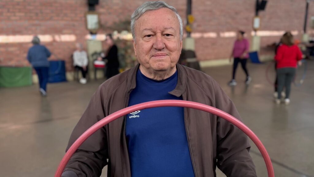 Um homem idoso sorri enquanto segura um bambolê rosa em primeiro plano, com outras pessoas praticando atividades físicas ao fundo.
A imagem mostra um homem mais velho, com cabelos grisalhos e pele enrugada, vestindo uma jaqueta marrom sobre uma camiseta azul escura com o logo da Umbro. Ele está de frente para a câmera, segurando um bambolê rosa que ocupa a parte inferior da imagem. Seu rosto exibe um sorriso gentil e ele parece relaxado e contente. O fundo da imagem está desfocado, mas é possível distinguir um ambiente interno que parece ser uma quadra ou ginásio, com paredes de tijolos aparentes e iluminação artificial. Há outras pessoas ao fundo, também em movimento, parecendo engajadas em alguma atividade física, como dança ou exercícios com bambolê, como sugerido pela presença de múltiplos bambolês e pelas posturas das pessoas. Uma mulher vestindo uma blusa rosa e calças escuras está à esquerda do centro, enquanto outra mulher de blusa vermelha e calças cinzas está mais à direita. Menos discernível, há uma pessoa sentada em uma cadeira ao fundo. A atmosfera geral da imagem é de um ambiente comunitário e ativo, focado em bem-estar e lazer para idosos, com um tom positivo e acolhedor. As cores predominantes são os tons terrosos da jaqueta e das paredes de tijolo, contrastando com o azul vibrante da camiseta e o rosa chamativo do bambolê. A composição coloca o homem em primeiro plano como foco principal, mas sugere a existência de uma atividade coletiva acontecendo em segundo plano.