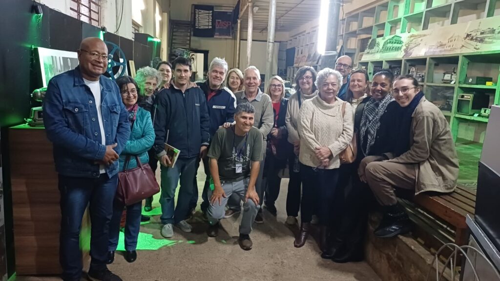 Foto de um grande grupo posando em ambiente interno com aparência de galpão, ateliê ou espaço cultural. Principais elementos: Pessoas: cerca de 16–18 pessoas de idades variadas (jovens, adultos e idosos) reunidas próximas umas das outras, muitas sorrindo. Um homem está agachado no centro-frente; à direita, duas pessoas sentadas/apoIadas em um banco de madeira; à esquerda, um homem de jaqueta jeans em primeiro plano. Roupas e acessórios: vestuário casual e camadas (casacos, suéteres, jaquetas), algumas bolsas e óculos; um crachá pendurado no pescoço do homem agachado, sugerindo organização ou participação formal no evento. Ambiente: espaço interno com prateleiras abertas ao fundo (alguns objetos/peças expostas), estruturas metálicas e vigas aparentes no teto. Paredes escuras à esquerda com luzes verdes que projetam iluminação colorida em alguns pontos. Iluminação: mistura de luz ambiente amarelada e pontos de luz verde que criam reflexos no piso; iluminação geral suficiente para enxergar detalhes dos rostos e do ambiente. Objetos e detalhes: à esquerda há um móvel ou balcão de madeira e equipamentos (possivelmente objetos de exposição); ao fundo, banners ou cartazes pendurados no alto; banco de madeira à direita e chão de piso cimentado. Atmosfera: sensação de encontro comunitário, visita guiada, oficina ou reunião cultural — clima amigável e informal.