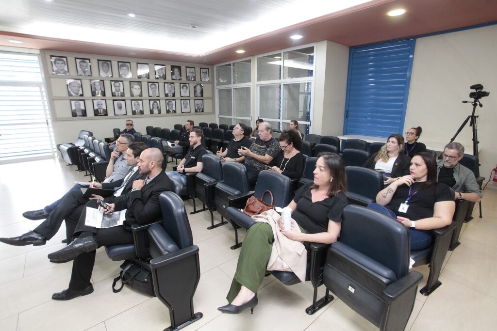 A imagem mostra uma reunião, palestra ou evento ocorrendo em um auditório ou sala de conferências. Aqui estão os detalhes principais: O Ambiente: É uma sala bem iluminada com fileiras de poltronas estofadas em azul escuro. Nas paredes, há uma galeria de fotos de pessoas, possivelmente retratos de autoridades ou homenageados anteriores. Os Participantes: Há um grupo de cerca de 15 pessoas, vestidas de forma casual-elegante ou formal (algumas de terno, outras de roupas casuais). A maioria está sentada, prestando atenção em algo à frente (provavelmente um palestrante ou apresentação que não aparece na foto). Algumas pessoas estão mexendo em seus celulares, enquanto outras observam atentamente. Equipamento: No canto direito da sala, há um tripé com uma câmera montada, indicando que o evento está sendo filmado ou transmitido. Atmosfera: O ambiente parece formal e profissional, típico de um espaço institucional.