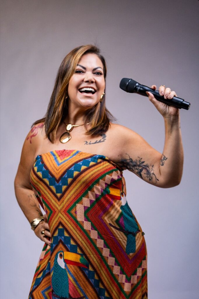 mulher sorridente segurando um microfone, com postura confiante — parece cantora ou apresentadora em sessão fotográfica ou performance. Pontos principais: Pessoa: mulher adulta, expressão alegre e confiante; rosto visível, não identificável. Ação: segurando um microfone na mão direita, posando como se estivesse prestes a falar ou cantar. Roupa: vestido colorido com padrão geométrico vibrante e ilustrações de tucanos. Detalhes: tatuagens visíveis no braço e no peito, colar e brincos dourados, unhas feitas e acessórios no pulso. Fundo e iluminação: fundo neutro e iluminação suave, foco principal na pessoa. Atmosfera: imagem transmite energia positiva, performance e estilo pessoal marcante.