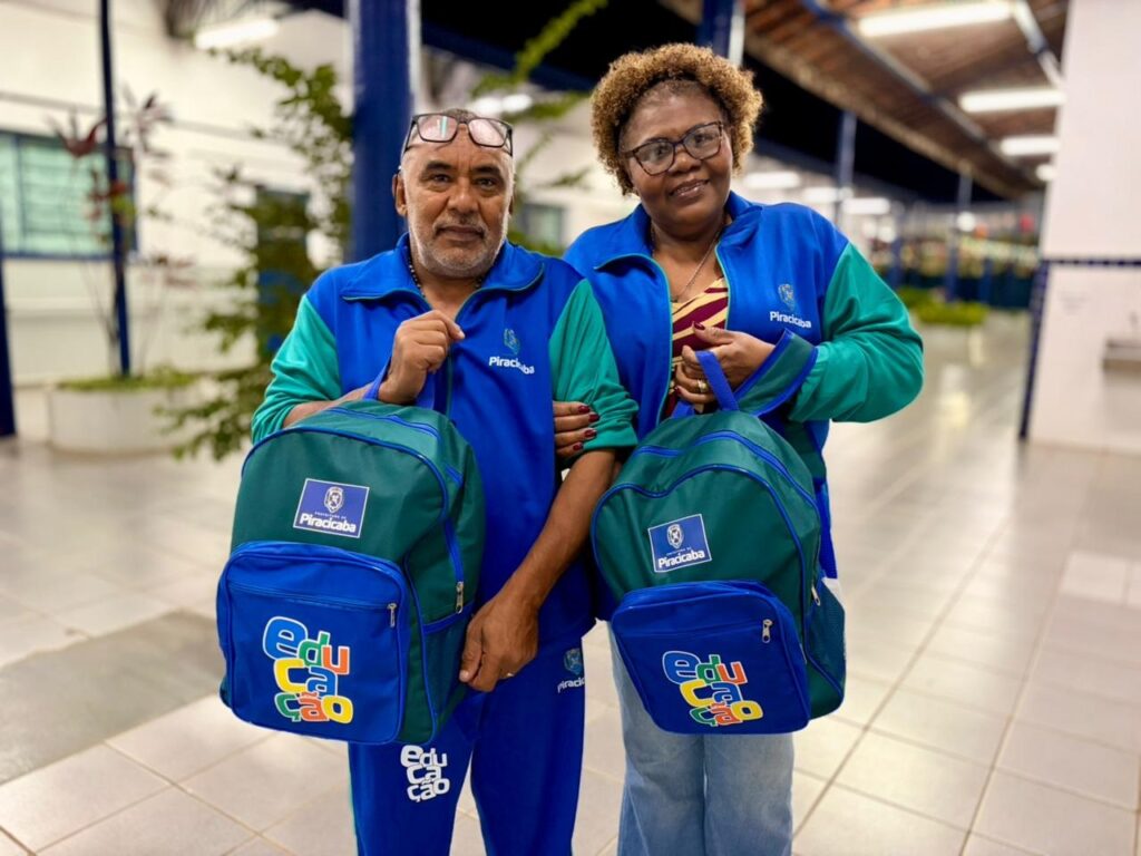 Dois adultos em pé, posando lado a lado num corredor iluminado (provavelmente institucional). Ambos seguram mochilas verdes e azuis com o logo "Piracicaba" e a palavra colorida "educação" no bolso frontal. Pessoas: homem à esquerda e mulher à direita, sorrindo levemente. O homem tem cabelo curto e barba grisalha; usa óculos apoiados na testa. A mulher usa óculos, cabelo cacheado curto e joias discretas (anel/colar). Roupa: ambos vestem agasalhos na paleta azul e verde com o emblema "Piracicaba" — parecem uniformes institucionais. O homem também usa calça azul com a palavra "educação" impressa. Mochilas: dois modelos idênticos, verdes com detalhes azuis; etiqueta com brasão/texto "Piracicaba" na parte superior e estampa colorida "educação" no bolso frontal. Alças e zíperes visíveis, material sintético resistente. Ambiente: piso de cerâmica clara, colunas e jardineiras ao fundo, iluminação forte de teto; sensação de corredor amplo e organizado, possivelmente dentro de escola ou centro público. Atmosfera: momento de entrega ou recebimento de material escolar — clima amistoso e institucional, com foco em educação/comunidade. Dois adultos em pé, posando lado a lado num corredor iluminado (provavelmente institucional). Ambos seguram mochilas verdes e azuis com o logo "Piracicaba" e a palavra colorida "educação" no bolso frontal. Pessoas: homem à esquerda e mulher à direita, sorrindo levemente. O homem tem cabelo curto e barba grisalha; usa óculos apoiados na testa. A mulher usa óculos, cabelo cacheado curto e joias discretas (anel/colar). Roupa: ambos vestem agasalhos na paleta azul e verde com o emblema "Piracicaba" — parecem uniformes institucionais. O homem também usa calça azul com a palavra "educação" impressa. Mochilas: dois modelos idênticos, verdes com detalhes azuis; etiqueta com brasão/texto "Piracicaba" na parte superior e estampa colorida "educação" no bolso frontal. Alças e zíperes visíveis, material sintético resistente. Ambiente: piso de cerâmica clara, colunas e jardineiras ao fundo, iluminação forte de teto; sensação de corredor amplo e organizado, possivelmente dentro de escola ou centro público. Atmosfera: momento de entrega ou recebimento de material escolar — clima amistoso e institucional, com foco em educação/comunidade. Dois adultos em pé, posando lado a lado num corredor iluminado (provavelmente institucional). Ambos seguram mochilas verdes e azuis com o logo "Piracicaba" e a palavra colorida "educação" no bolso frontal. Pessoas: homem à esquerda e mulher à direita, sorrindo levemente. O homem tem cabelo curto e barba grisalha; usa óculos apoiados na testa. A mulher usa óculos, cabelo cacheado curto e joias discretas (anel/colar). Roupa: ambos vestem agasalhos na paleta azul e verde com o emblema "Piracicaba" — parecem uniformes institucionais. O homem também usa calça azul com a palavra "educação" impressa. Mochilas: dois modelos idênticos, verdes com detalhes azuis; etiqueta com brasão/texto "Piracicaba" na parte superior e estampa colorida "educação" no bolso frontal. Alças e zíperes visíveis, material sintético resistente. Ambiente: piso de cerâmica clara, colunas e jardineiras ao fundo, iluminação forte de teto; sensação de corredor amplo e organizado, possivelmente dentro de escola ou centro público. Atmosfera: momento de entrega ou recebimento de material escolar — clima amistoso e institucional, com foco em educação/comunidade.
