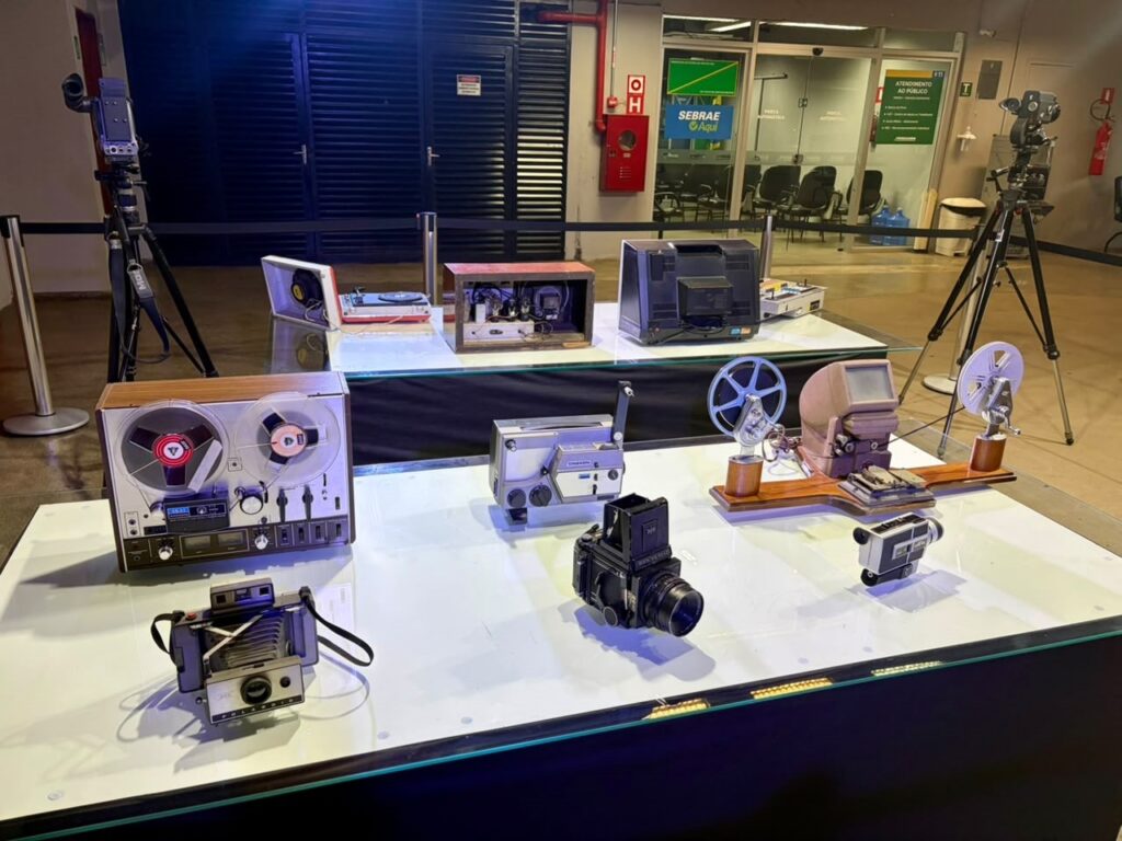 Esta foto mostra uma exposição fascinante de equipamentos audiovisuais antigos, organizados sobre mesas brancas iluminadas em um ambiente que parece ser um centro cultural ou museu. É um verdadeiro mergulho na história da tecnologia! Aqui está o que podemos ver, detalhe por detalhe: Mesa da frente (em primeiro plano): Esquerda: Um gravador de rolo (fita magnética) da marca Akai, um clássico do som analógico de alta fidelidade. Ao lado dele, uma câmera Polaroid instantânea antiga, com seu fole característico. Centro: Uma câmera fotográfica de formato médio (parece uma Pentacon Six ou similar) e, ao lado dela, um projetor de filmes pequeno, provavelmente de 8mm ou Super 8. Direita: Um equipamento de edição de filme com rolos (moviolinha) e uma câmera de filmagem menor, também de 8mm. Mesa de trás: Esquerda: Um rádio antigo aberto, revelando seus componentes internos (válvulas e fios), e um toca-discos de vinil portátil. Direita: Um monitor de vídeo (televisor de tubo) antigo, posicionado de costas, e mais alguns equipamentos de áudio/vídeo menores ao lado. Ao redor: Há dois tripés profissionais nas laterais da foto, segurando câmeras de cinema e vídeo antigas, como se estivessem prontas para gravar. O fundo mostra uma estrutura de prédio com uma porta de vidro, onde se vê uma placa do "Sebrae Aqui", sugerindo que a exposição está em um espaço público ou centro comercial. É uma coleção nostálgica incrível para quem ama fotografia e cinema!
