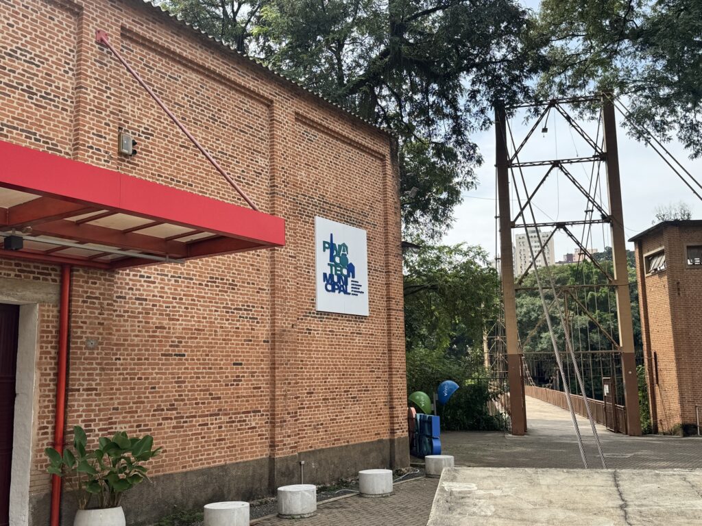 Imagem de área externa com parede de tijolos aparentes e uma ponte suspensa ao lado direito. Fachada: parede de tijolos vermelhos com detalhes em relevo; beiral com telha ondulada e estrutura metálica vermelha formando uma pequena marquise sobre a entrada. Elementos visuais: cano vertical vermelho na parede, luminária externa, e um letreiro branco afixado na fachada com logotipo azul e texto (aparentemente indicando um espaço municipal ou cultural). Acessórios de chão: vasos brancos com plantas na base da parede e blocos cilíndricos de concreto formando limite/piso. Ponte: estrutura metálica alta e pórtico com cabos de suspensão visíveis que levam a uma passarela de madeira; a ponte conecta a área da direita a um outro ponto mais abaixo. Vegetação e entorno: árvores altas ao fundo por cima da parede e vegetação densa além da ponte; ao longe, prédios urbanos visíveis entre as copas. Iluminação e clima: luz natural clara, céu parcialmente nublado; sombras suaves indicando dia claro. Atmosfera: local parece ser parte de um equipamento público (cultural ou municipal) integrado à paisagem, com acessibilidade pela ponte e acabamento rústico em tijolo.