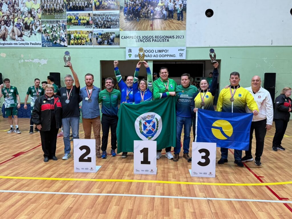 Esta é uma foto comemorativa de uma premiação esportiva, tirada em uma quadra poliesportiva de madeira. O cenário registra o pódio dos "Jogos Regionais 2023" realizados em Lençóis Paulista. Aqui estão os detalhes da cena: O Pódio: Há três placas numeradas no chão (2, 1 e 3), indicando as posições. As Pessoas: Um grupo grande de pessoas está posando junto. Muitos vestem camisas de uniforme esportivo (tons de verde, azul e amarelo) e medalhas no pescoço. Três pessoas seguram troféus dourados erguidos. As Bandeiras: No centro, o grupo segura uma bandeira verde com o brasão de um município. À direita, uma pessoa segura uma bandeira azul com um símbolo circular amarelo, representando a delegação ou equipe. O Fundo: Há uma grande faixa na parede ao fundo com colagens de fotos de edições anteriores dos jogos (2018 a 2023) e os dizeres "CAMPEÕES JOGOS REGIONAIS 2023 LENÇÓIS PAULISTA" e "JOGO LIMPO! DIGA NÃO AO DOPING". O Ambiente: Além dos premiados, há outras pessoas nas laterais (algumas vestindo camisas de times, outras com agasalhos), sugerindo um evento organizado com vários participantes e equipe de apoio. É uma imagem que transmite espírito esportivo, conquista e celebração de uma competição regional.