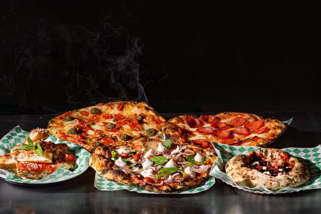 Esta é uma imagem apetitosa de uma seleção de pizzas recém-saídas do forno, dispostas sobre uma superfície escura. Aqui estão os detalhes do que podemos ver: Pizzas principais: Há várias pizzas com diferentes coberturas, todas com bordas levemente tostadas, indicando um forno a lenha. No topo à esquerda, uma pizza com molho de tomate, tomates cereja e o que parece ser pesto ou ervas. É desta pizza que se vê uma fumaça clara subindo, sugerindo que ela está bem quente. Ao lado, à direita, uma clássica pizza de pepperoni, com as fatias de salame bem distribuídas. Na parte inferior central, uma pizza mais elaborada, com cogumelos, cebola roxa, vegetais e pequenas porções de queijo branco (como ricota ou queijo de cabra), decorada com folhas frescas. Acompanhamento: À esquerda, há um prato menor com pedaços de massa de pizza ou focaccia, cobertos com molho, queijo e ervas. Sobremesa/Outro: À direita, há um item menor, de formato circular, que parece ser uma mini pizza doce ou uma massa recheada com calda escura (talvez chocolate ou redução de frutas vermelhas) e decorada com flores comestíveis. Apresentação: Tudo está servido sobre papéis quadriculados verdes e brancos, criando um contraste visual interessante. A iluminação é dramática e focada, destacando a textura e o calor das massas.