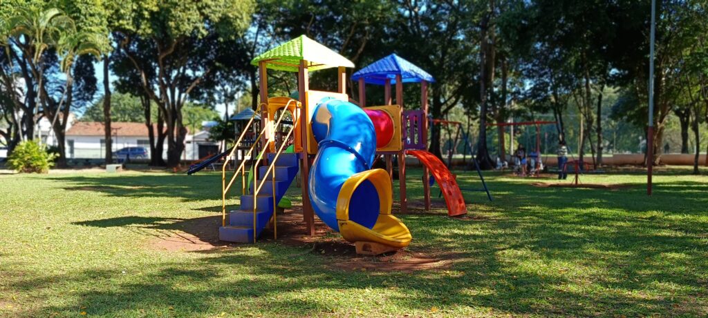 Esta imagem mostra um parquinho infantil colorido situado em uma área verde arborizada, sob um dia ensolarado. Aqui estão os detalhes principais: O Equipamento: No centro, há um brinquedo de playground modular grande, feito de plástico vibrante, com tons de azul, amarelo, vermelho e verde. Ele possui escadas de acesso, um escorregador em espiral (azul e amarelo) e uma plataforma coberta por um telhado verde. O Ambiente: O parquinho está instalado em um gramado aberto. Ao redor, há muitas árvores altas que criam um cenário sombreado e tranquilo. Detalhes Adicionais: Ao fundo, é possível ver outros equipamentos de recreação, como balanços e talvez um carrossel, além de algumas pessoas ao longe curtindo o local. Clima: A luz do sol está bem forte, criando um contraste claro entre as áreas iluminadas e as sombras das árvores projetadas no chão, sugerindo um dia agradável e bom para brincar ao ar livre.