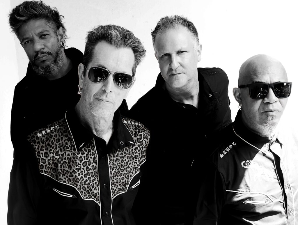 Esta foto em preto e branco mostra quatro homens posando juntos, com uma estética que lembra uma banda de rock. Eles estão com expressões sérias e olhar direto para a câmera. O primeiro plano: À esquerda, um homem com óculos escuros usa uma camisa com detalhes em estampa de leopardo no peito e a palavra "SABOT" bordada. À direita, um homem careca também de óculos escuros usa uma camisa preta com detalhes bordados semelhantes. O fundo: Atrás deles, há outros dois homens. Um à esquerda com barba e cabelo estilizado, e outro à direita com um semblante mais neutro e camisa preta lisa. Todos transmitem uma atitude marcante e rock and roll!