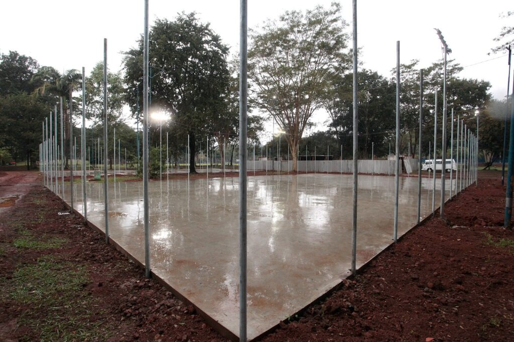 Essa imagem mostra uma quadra esportiva, provavelmente de futsal ou poliesportiva, que parece estar em construção ou reforma. Aqui estão os detalhes principais: Piso: O chão é de cimento liso e está bem molhado, como se tivesse chovido recentemente. Estrutura: Vários postes metálicos altos estão instalados ao redor de todo o perímetro da quadra, sugerindo que uma grade ou rede de proteção será colocada ali. Ambiente: A quadra está cercada por árvores e vegetação, indicando que fica em uma área aberta, como um parque ou praça. Iluminação: Há holofotes acesos que brilham sobre a superfície molhada do piso. Ao fundo, dá para ver uma pequena cerca de proteção e um veículo utilitário branco estacionado.