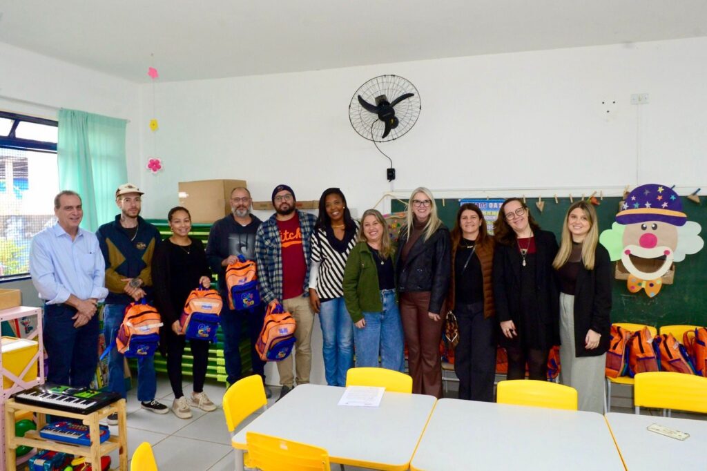 Foto interna de uma sala de aula infantil com um grupo de 12 adultos alinhados de pé em frente ao quadro-negro, posando para a foto. Pessoas: 12 adultos (homens e mulheres) lado a lado, sorrindo; alguns seguram mochilas escolares laranja e azul. Não faço identificação pessoal. Mobiliário: mesas e cadeiras infantis amarelas e brancas em primeiro plano; estante pequena com brinquedos e teclados à esquerda. Material escolar: várias mochilas laranja e azul empilhadas sobre cadeiras e no chão à direita; caixas e materiais empilhados ao fundo. Decoração e ambiente: quadro-negro atrás do grupo com cartazes e varalzinho de pregadores; grande figura decorativa de palhaço colorido fixada à direita do quadro. Estrutura: parede branca, janela com cortina verde clara à esquerda, ventilador de parede acima do grupo e caixas de papelão empilhadas ao fundo. Iluminação e clima: luz natural entrando pela janela, ambiente claro e organizado, típica sala de educação infantil em evento de entrega de materiais ou visita institucional.