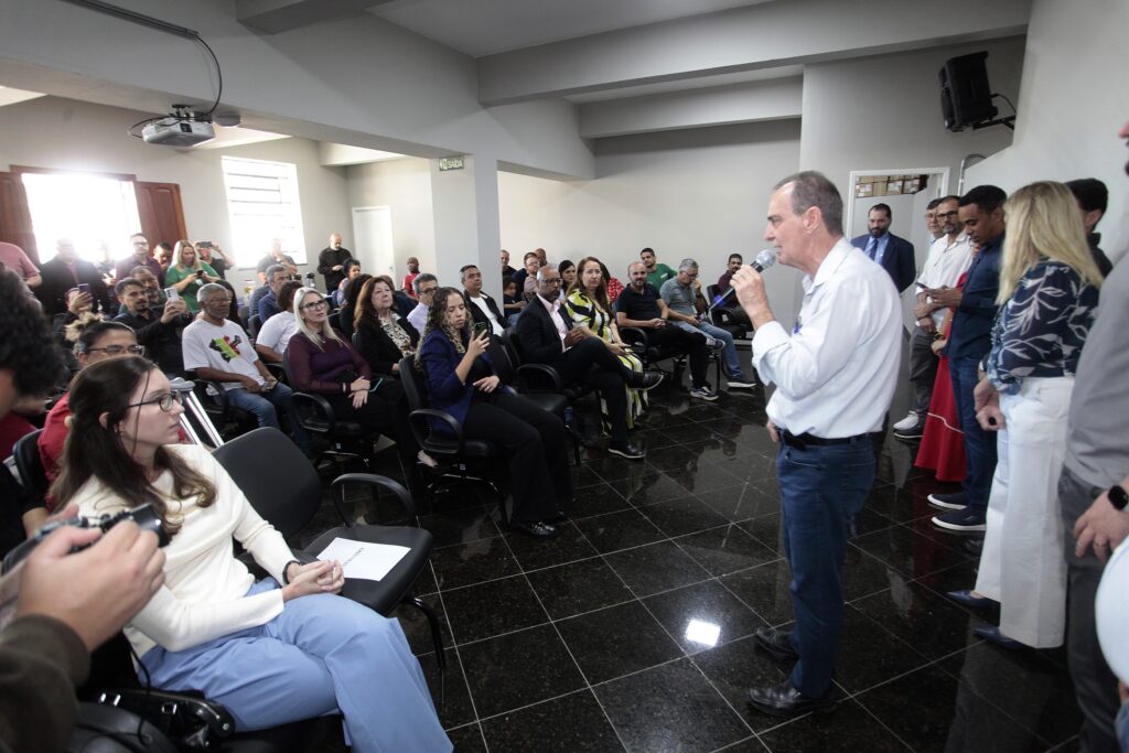 Esta imagem retrata uma reunião ou apresentação em um ambiente corporativo ou institucional. Palestrante: No lado direito da foto, um homem de camisa social azul clara e calça jeans escura fala ao microfone, dirigindo-se ao público presente. Plateia: Várias pessoas estão sentadas em cadeiras de escritório pretas, ouvindo atentamente. Entre o público, nota-se uma mulher em primeiro plano, à esquerda, com blusa branca de gola alta e calça azul, olhando para o palestrante, e outras pessoas logo atrás segurando celulares ou acompanhando a fala. Ambiente: O local possui piso de cerâmica preta brilhante e paredes em tons neutros. Há um grupo de pessoas em pé no lado direito, perto do palestrante, que parecem ser organizadores ou convidados especiais. O ambiente é bem iluminado, com um projetor fixado no teto e uma placa indicando "SAÍDA" ao fundo.