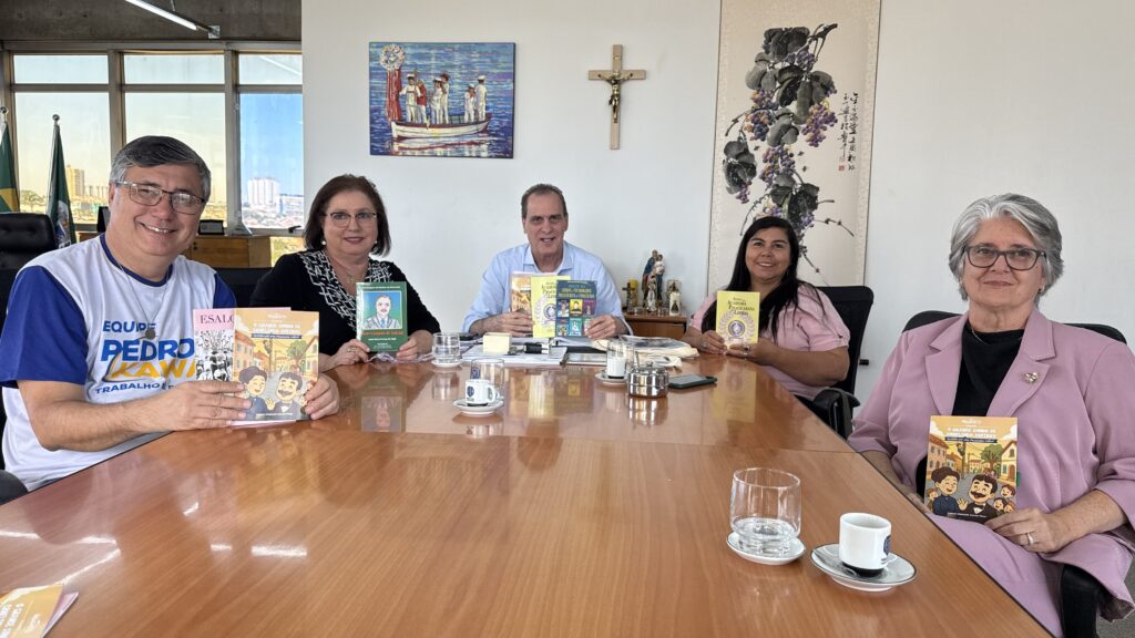 Foto horizontal de uma reunião informal em uma sala de escritório/gabinete, com cinco pessoas sentadas lado a lado atrás de uma mesa grande de madeira clara. Elas seguram livros ou folhetos coloridos, posando para a foto e sorrindo. Detalhes visuais: Pessoas: cinco adultos (três mulheres e dois homens) dispostos em linha. Todos sorriem e olham para a câmera. Não é possível identificar quem são. À esquerda: homem com óculos e camiseta branca com mangas azuis, segurando um livro infantil com ilustrações na capa. Próxima a ele: mulher de cabelos curtos castanhos, óculos, roupa escura com padrão claro, também segura um livreto. No centro: homem de camisa clara, segurando dois ou três exemplares empilhados de publicações. À direita do centro: mulher de cabelos longos e escuros, blusa rosa clara, segura um livreto amarelo. Extremidade direita: mulher de cabelo grisalho curto, óculos e blazer rosa claro, também com um livro nas mãos. Mesa: superfície envidraçada/cloth refletiva com xícaras de café, pires, copos de água, cinzeiro metálico, telefone, papéis e pequenos objetos espalhados. Fundo e decoração: grandes janelas à esquerda com vista parcial da cidade; bandeiras ao fundo (à esquerda). Na parede atrás das pessoas há um crucifixo em madeira, um quadro colorido de uma cena náutica e um painel vertical com ilustração de uvas e escrita oriental. Também há pequenas imagens religiosas sobre um móvel no fundo. Iluminação: natural, vinda das janelas; ambiente claro e acolhedor. Tom e atmosfera: encontro institucional ou cultural, descontraído e cordial, possivelmente para apresentação ou lançamento de publicações. Os livros/folhetos têm capas ilustradas e coloridas, sugerindo materiais voltados a público geral ou infantojuvenil.