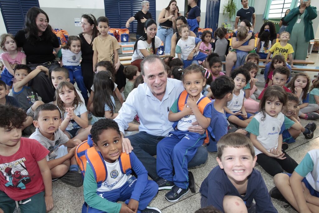Uma grande foto de grupo em ambiente escolar mostrando muitas crianças pequenas sentadas no chão, com alguns adultos em pé ou agachados entre elas. A cena é alegre e movimentada. Centrado-em-praimeiro-plano: um homem adulto agachado sorrindo — ele segura no colo uma menina pequena que sorri amplamente. Ao lado, outro menino e outra menina, ambos com mochilas laranja e uniformes escolares azul/verde com o nome “Piracicaba” visível. Crianças: muitas crianças pré‑escolares sentadas no piso, em diferentes posturas (algumas cruzando as pernas, outras apoiadas nas mãos). Expressões variam: sorrisos, olhares curiosos, distração. Há diversidade de tons de pele e cabelos (lisos, cacheados). Adultos: várias mulheres e homens ao fundo e nas laterais — algumas sentadas em bancos, outras em pé observando, algumas sorrindo, outras conversando. Uma mulher à esquerda sorri olhando para as crianças; outra à direita filma ou fotografa com um celular. Ambiente: sala ampla com piso de granito/cerâmica, bancos de madeira ao fundo e mochilas laranja empilhadas sobre os bancos. Paredes claras e janelas com venezianas azul‑escuro. Placas e material escolar ao fundo indicam espaço institucional/educacional. Vestuário e objetos: muitos uniformes escolares brancos e verdes com logo, mochilas alaranjadas, roupas casuais dos adultos (camisas, calças). Alguns acessórios como anéis, crachás e óculos aparecem.
