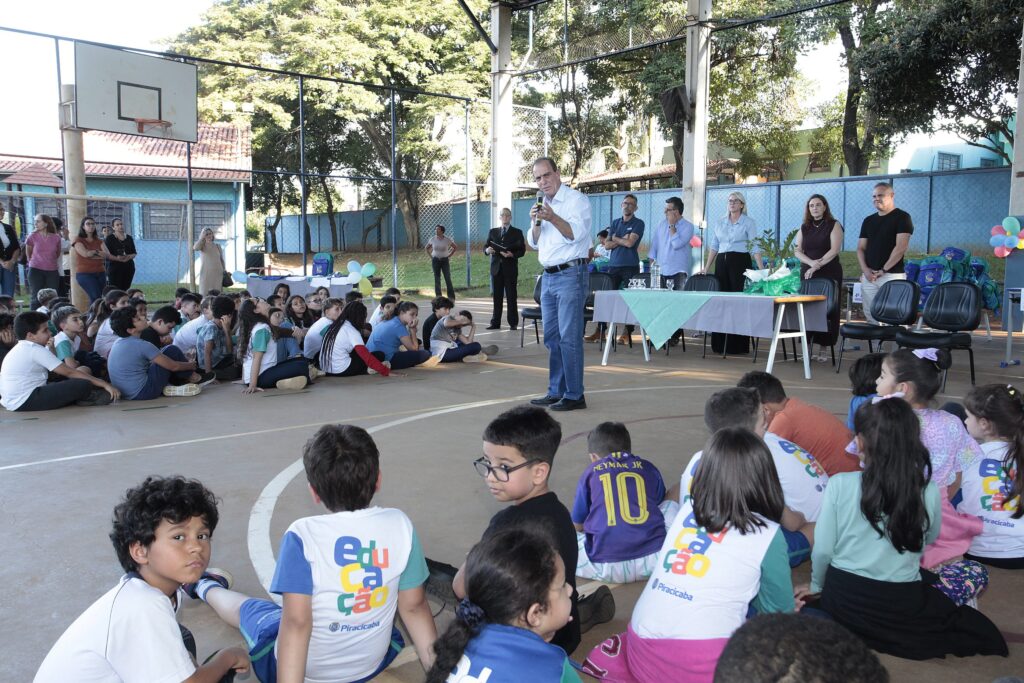 Foto de um evento escolar realizado em uma quadra coberta. Cena principal: muitas crianças sentadas no chão em grupos, viradas para a frente onde um homem em pé segura um microfone e fala. Mesa de honra: à direita há uma mesa com toalha, garrafas de água e cadeiras; atrás da mesa estão várias pessoas em pé (prováveis organizadores ou autoridades). Ambiente: quadra poliesportiva com tabela de basquete à esquerda, piso demarcado, grades laterais e árvores visíveis além da cerca. Elementos decorativos/infraestrutura: mochilas empilhadas e alguns balões coloridos junto à área dos organizadores; som ambiente aparente (caixa de som presa à coluna). Público: alunos usando uniformes escolares (alguns com a palavra “educação” nas camisetas) atentos ao discurso; também há adultos em pé ao fundo observando. Iluminação e clima: luz natural entrando pela lateral, ambiente ao ar livre parcialmente coberto, dia claro.