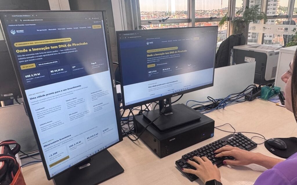 Foto de uma pessoa usando um computador em uma mesa de escritório, com dois monitores visíveis e janela grande ao fundo. Pessoa: apenas parte lateral da cabeça e as mãos aparecem; veste blusa roxa e usa relógio de pulso; não é possível identificar quem é. Monitores: dois monitores estilo paisagem lado a lado. O da esquerda está em modo retrato (vertical) mostrando um site com fundo escuro e texto em destaque amarelo: “Onde a inovação tem DNA de Piracicaba”. O monitor central mostra a mesma página em modo horizontal. Equipamento: teclado e mouse pretos sobre a mesa; CPU ou mini-PC preto apoiado sob o monitor central; vários cabos visíveis atrás do equipamento. Mesa e acessórios: bancada clara, mousepad preto circular, caneca/estojo listrado à esquerda e pequenos organizadores/verdinhos plásticos ao fundo à direita. Ambiente: janela larga ao fundo com vista urbana (prédios e telhados), plantas perto da janela e outros móveis/equipamentos de escritório ao redor. Iluminação: luz natural entrando pela janela, cena bem iluminada e com cores neutras. Impressão geral: ambiente de trabalho moderno, provavelmente alguém navegando ou conferindo conteúdo do site institucional de promoção/investimento de Piracicaba.