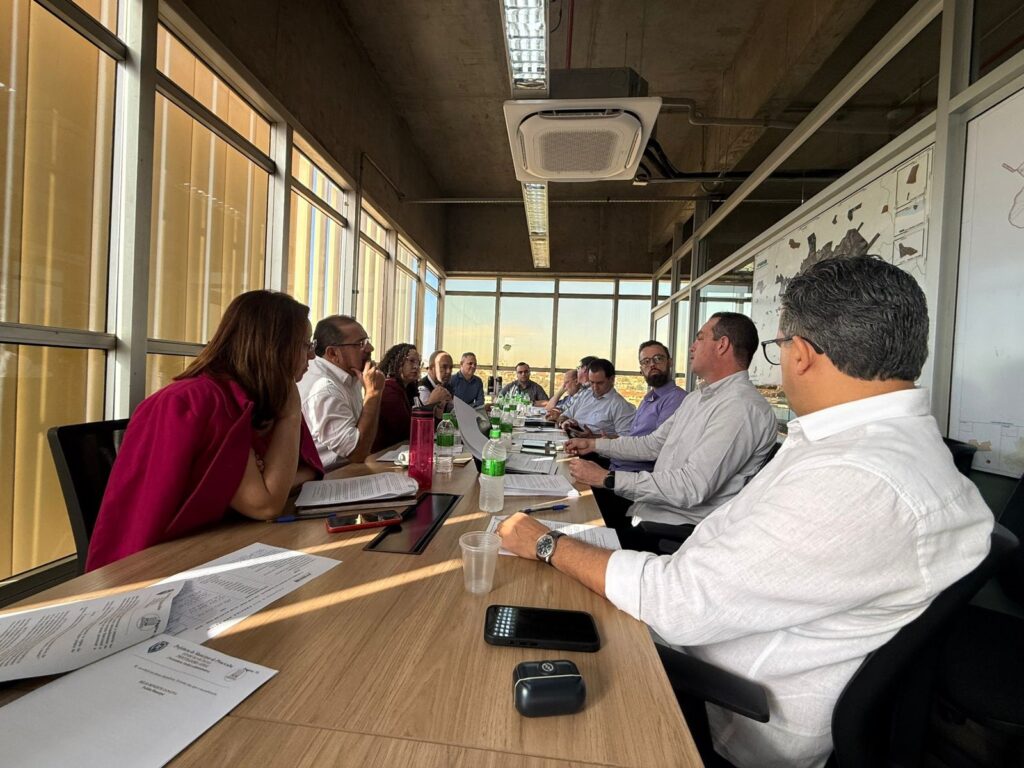 reunião presencial em sala de reunião com cerca de 10–12 pessoas sentadas ao redor de uma mesa comprida, discutindo documentos. Pontos principais: Ambiente: sala de reunião com paredes de vidro e janelas grandes, vista urbana e luz natural entrando pelo lado esquerdo. Participantes: várias pessoas (homens e mulheres) alinhadas dos dois lados da mesa, todas atentas à conversa; rostos não identificáveis. Mesa: documentos, folhas, canetas, garrafas de água e copos descartáveis espalhados; alguns celulares e um laptop aberto. Tom: encontro formal/institucional, discussão ou tomada de decisões ao fim da tarde (luz do sol baixa).