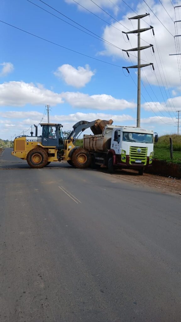 A imagem mostra uma operação de trabalho na beira de uma estrada asfaltada. Uma pá carregadeira amarela da marca John Deere (modelo 524K) está descarregando terra em uma caçamba de um caminhão basculante branco e verde da marca Volkswagen, que pertence à empresa "EcoTerra". O cenário é um dia ensolarado com céu azul e algumas nuvens. Há postes de rede elétrica percorrendo a lateral da via, em uma área que parece rural ou de obras de infraestrutura.