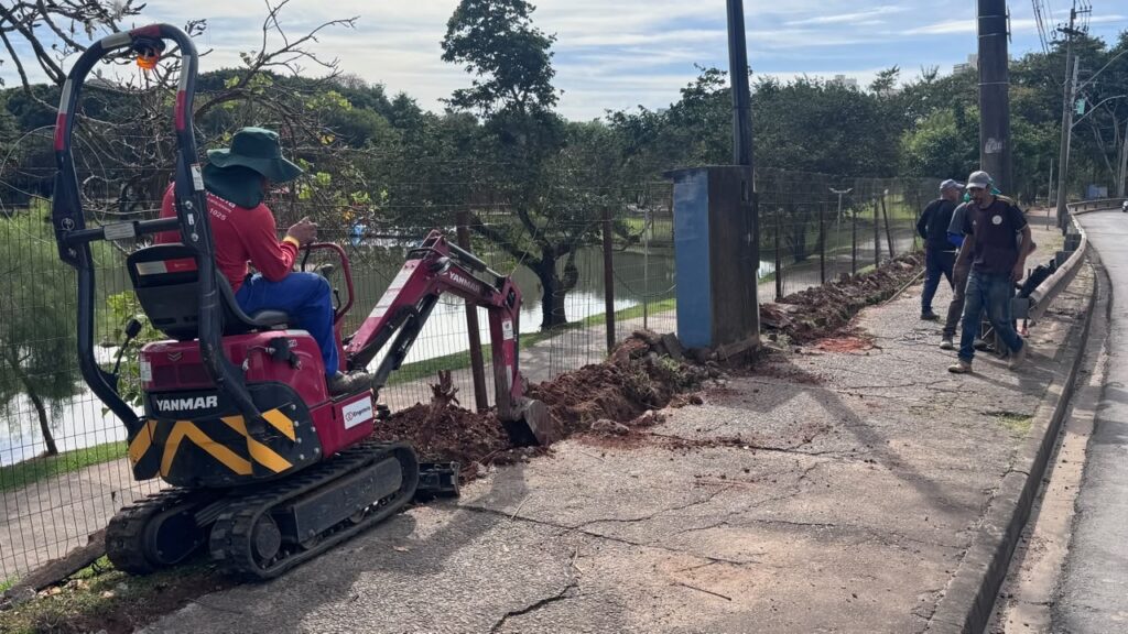 Nesta imagem, vemos um grupo de trabalhadores realizando uma obra de manutenção ou construção em uma calçada, ao lado de uma cerca que separa o caminho de uma área verde com um lago. Aqui estão os detalhes principais: Mini-escavadora: Em primeiro plano, uma escavadeira compacta da marca Yanmar, de cor vermelha, está sendo usada para abrir uma vala no chão, rente à cerca. Operador: Uma pessoa está sentada na escavadeira operando o equipamento. Ela usa uma camiseta vermelha, calça azul e um chapéu verde de proteção solar. Outros trabalhadores: Ao fundo, à direita, há dois outros homens caminhando pela calçada. Um deles usa uma camiseta preta e jeans, enquanto o outro, um pouco mais atrás, veste uma camisa escura. Ambiente: A cena acontece em uma área externa, possivelmente um parque ou espaço público urbano. Há uma grade metálica separando a calçada da vegetação e do espelho d'água ao fundo. O tempo parece estar claro e ensolarado.