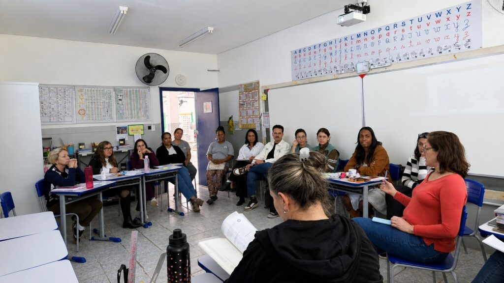 A imagem mostra uma reunião ou curso ocorrendo dentro de uma sala de aula. Há um grupo de cerca de 13 pessoas sentadas em mesas escolares, organizadas em formato de semicírculo, voltadas para uma mulher que está em pé, à direita, aparentemente conduzindo a atividade. A maioria dos participantes está concentrada, olhando para a palestrante ou para os materiais sobre as mesas. No centro, de costas, vê-se uma pessoa lendo um livro ou apostila. O ambiente é uma sala de aula comum: Há uma lousa branca grande ao fundo, com um cartaz acima exibindo o alfabeto em letras maiúsculas e minúsculas, além de ilustrações da linguagem de sinais (LIBRAS). Há cartazes informativos colados nas paredes laterais. Existe um ventilador de parede à esquerda e uma porta aberta que dá para um corredor. O piso é de cerâmica clara e a iluminação vem de luminárias fluorescentes no teto.